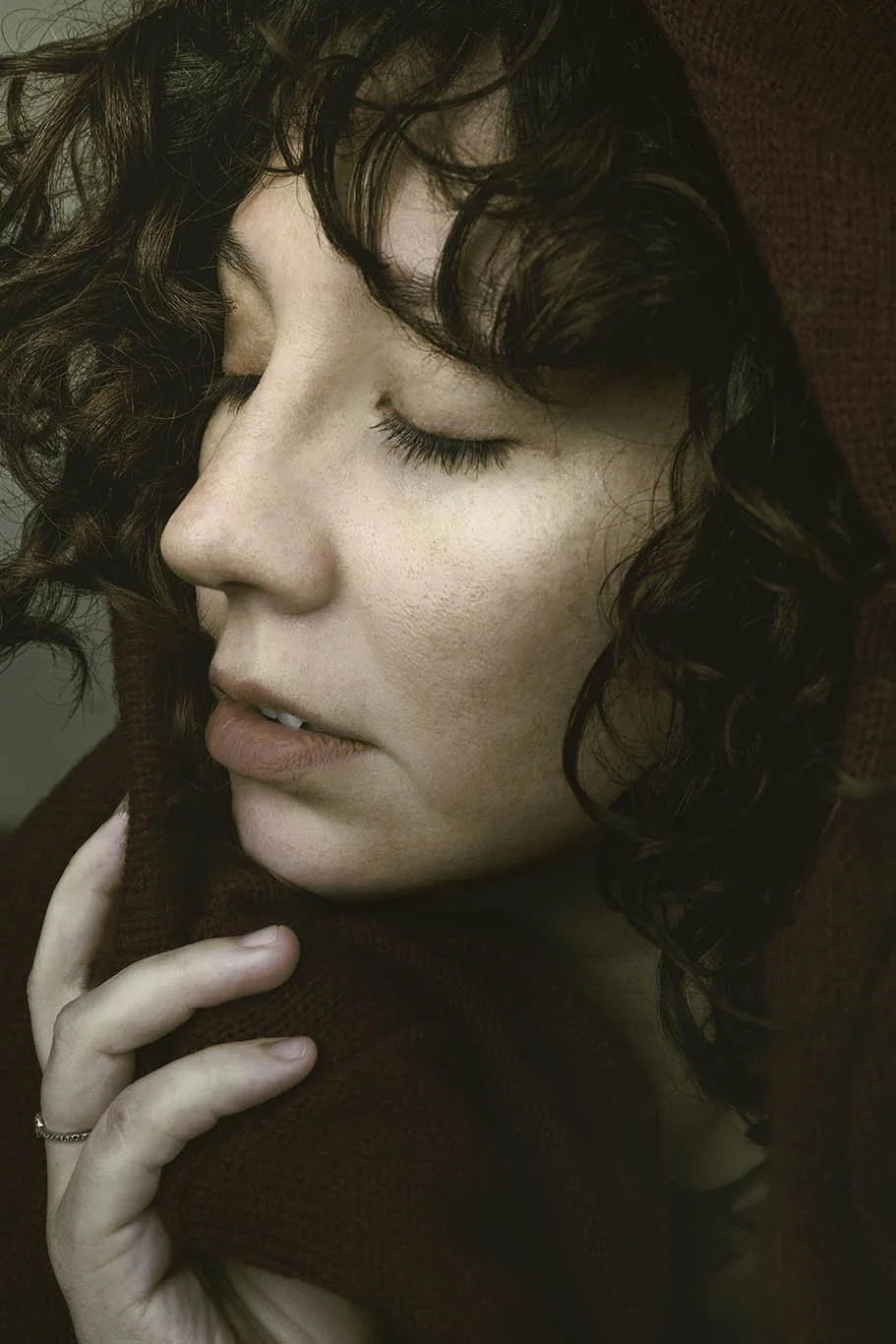 Close-up of a woman with dark curly hair and freckles, eyes closed, wearing a maroon hoodie.