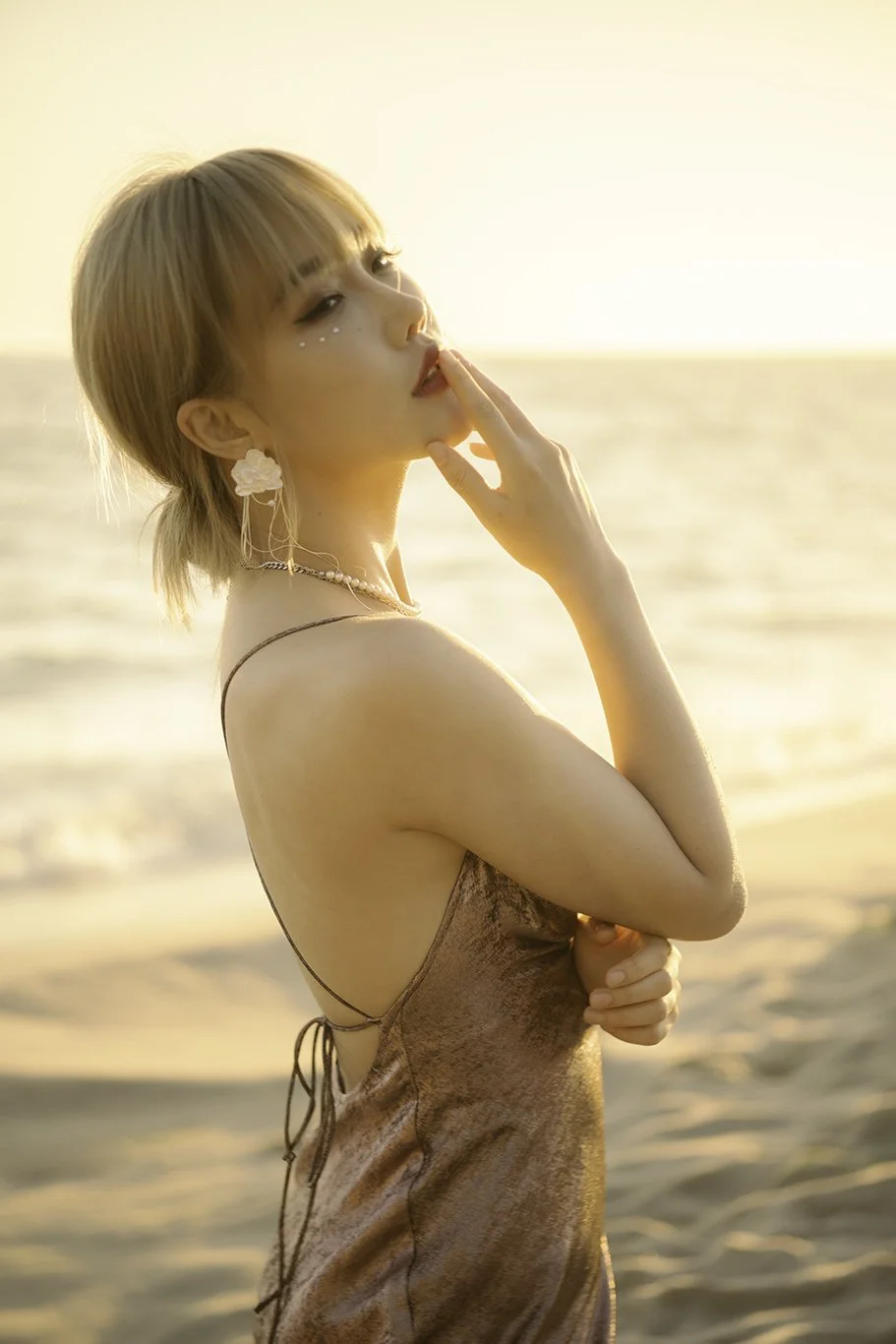 Woman with short blonde hair on a beach at sunset, touching her lips and looking into the distance.