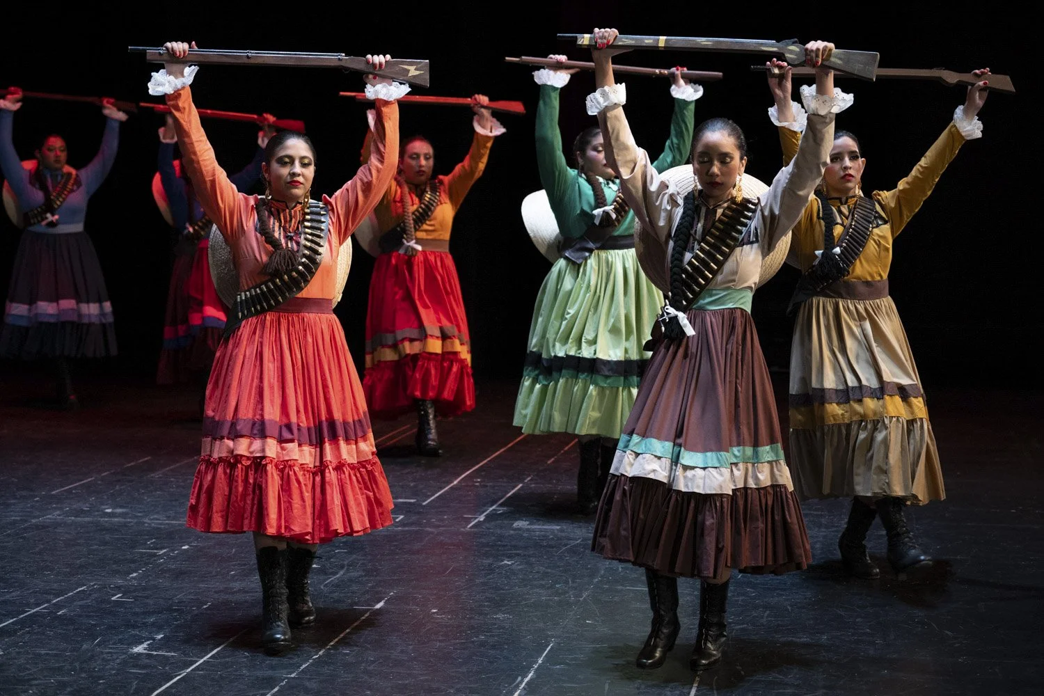 A group of women performing a traditional Mexican folk dance on a stage. They are wearing colorful, long, ruffled skirts and blouses, with some holding rifles pointed upward, and others wearing sombreros. The stage has a dark background.