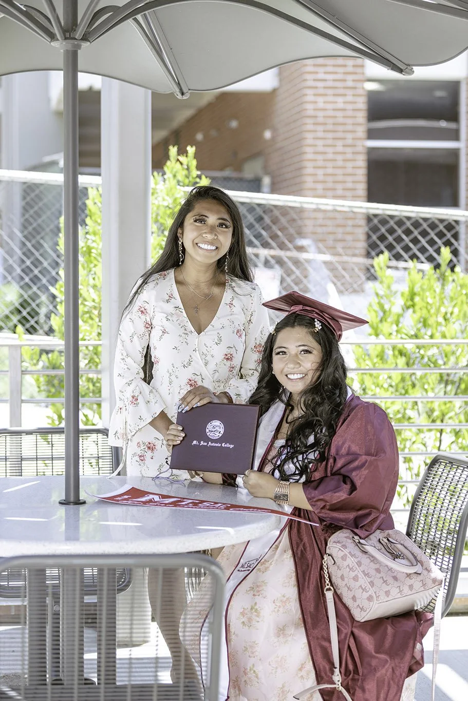 A young woman in a maroon graduation gown and cap holding a diploma, sitting at a table outdoors with another woman standing beside her, both smiling.