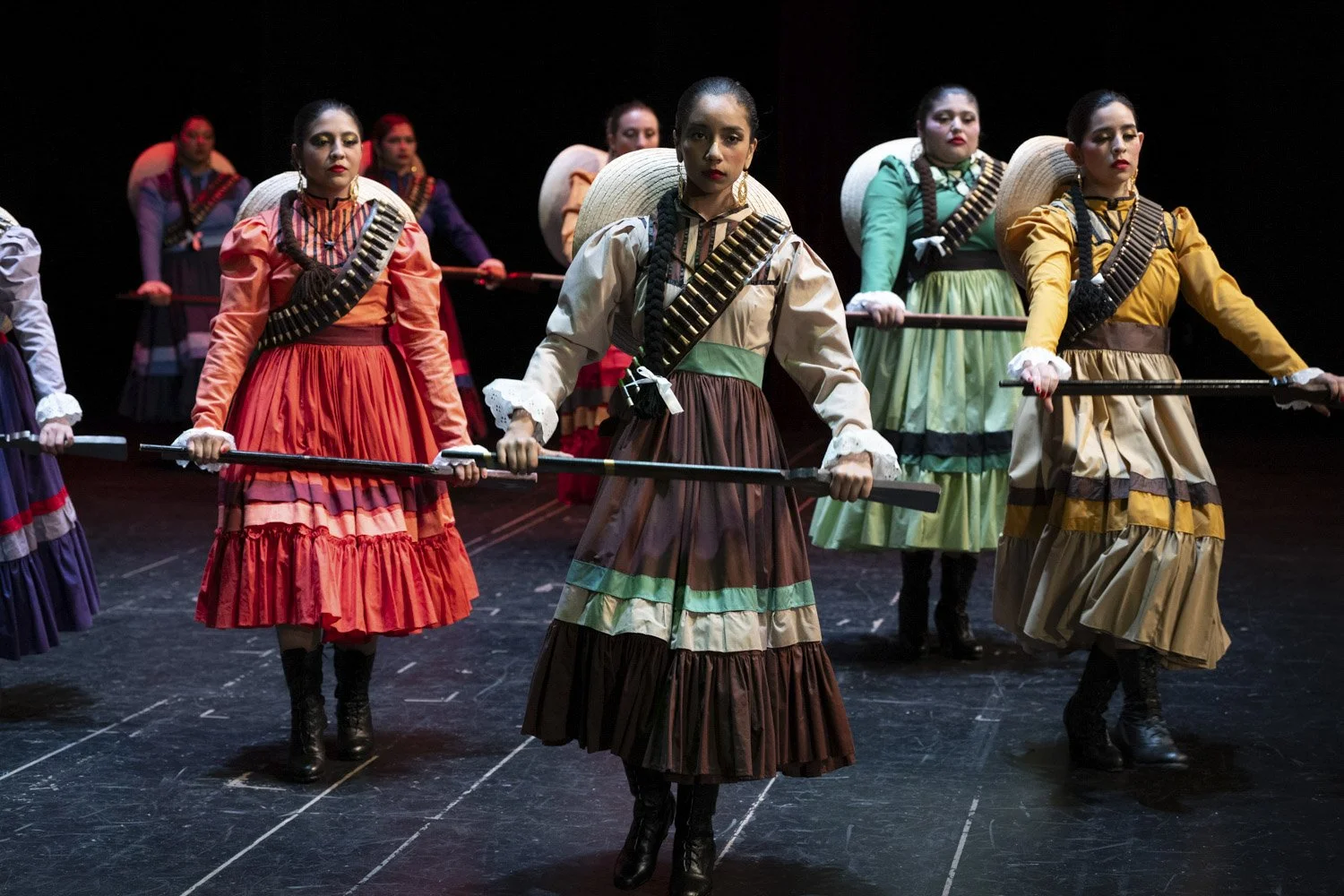 Women in traditional Mexican folkloric dresses holding sticks during performance on stage.