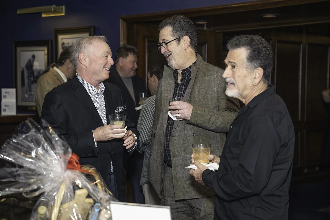Three men chatting and holding drinks at a social gathering in a room with dark wood paneled walls and framed black-and-white photographs.