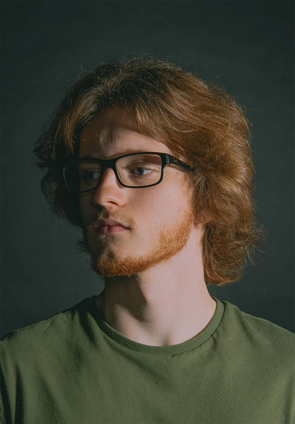 A young man with glasses, red hair, and a beard wearing a green shirt, looking off to the side against a dark background.