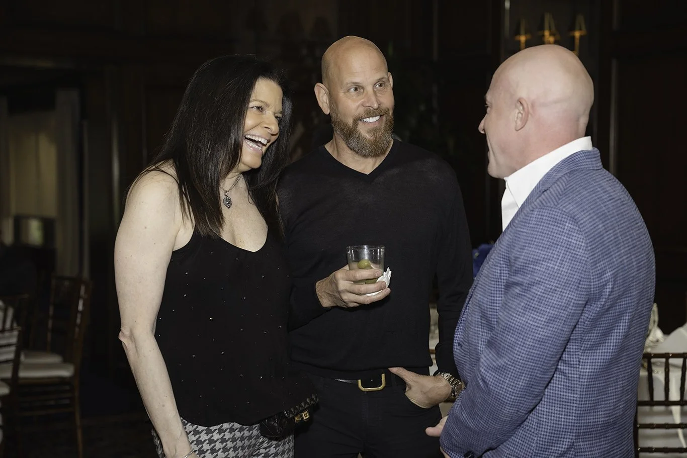 Three people engaged in conversation at a social event; two men and one woman are smiling and laughing, with the woman wearing a black dress and the man in the middle holding a drink.