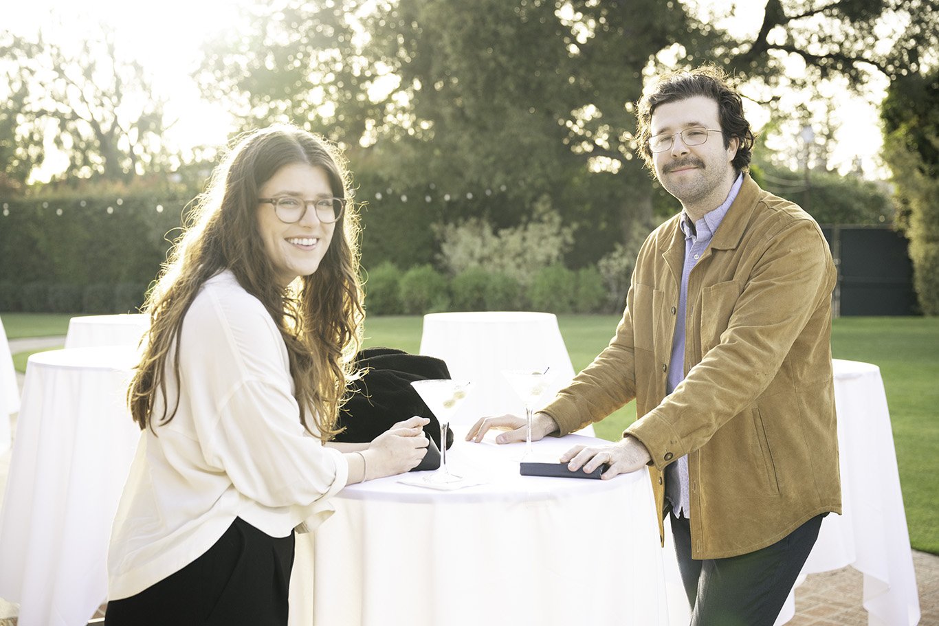A young woman and a young man are smiling outdoors at a social gathering, standing at a tall white cocktail table with drinks in glasses. The woman has long wavy hair and glasses, and the man has curly hair, glasses, and a mustache, both dressed casu