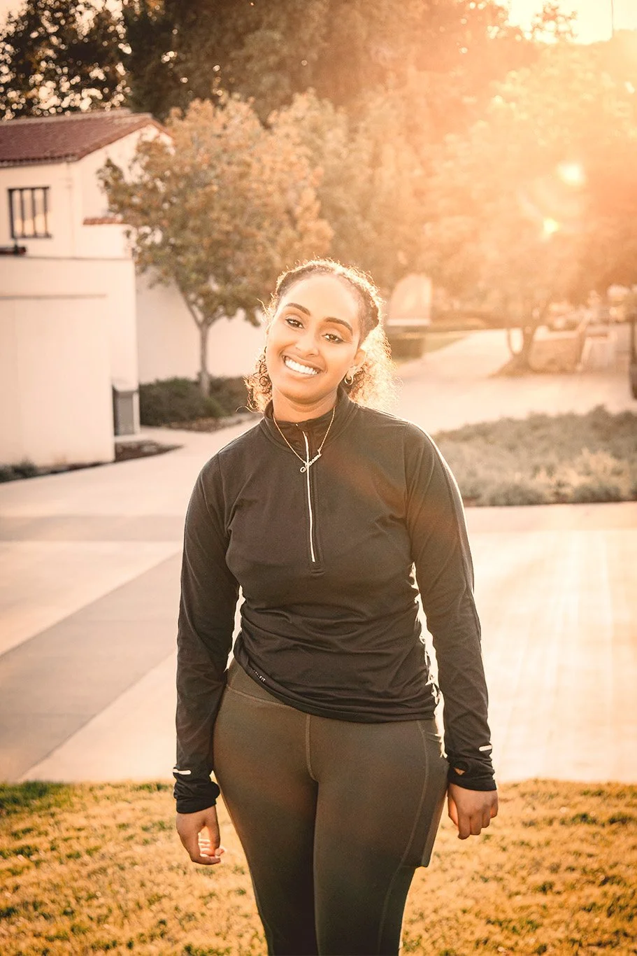 Smiling woman in athletic clothing standing outdoors during sunset in a residential area.