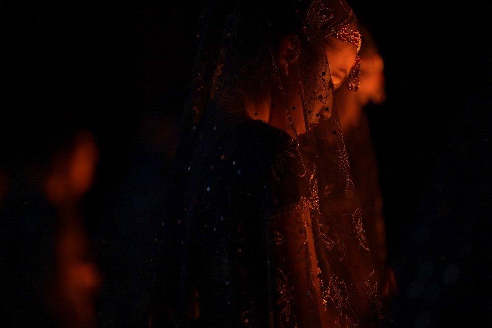 Woman dressed in ornate traditional attire with matching head covering illuminated by warm reddish light against dark background.