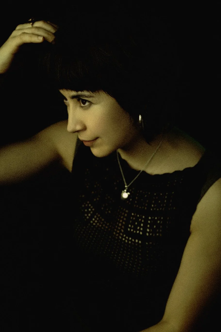 Woman with short dark hair wearing black top with cut-out pattern, necklace, and hoop earring touching her hair in dark environment.