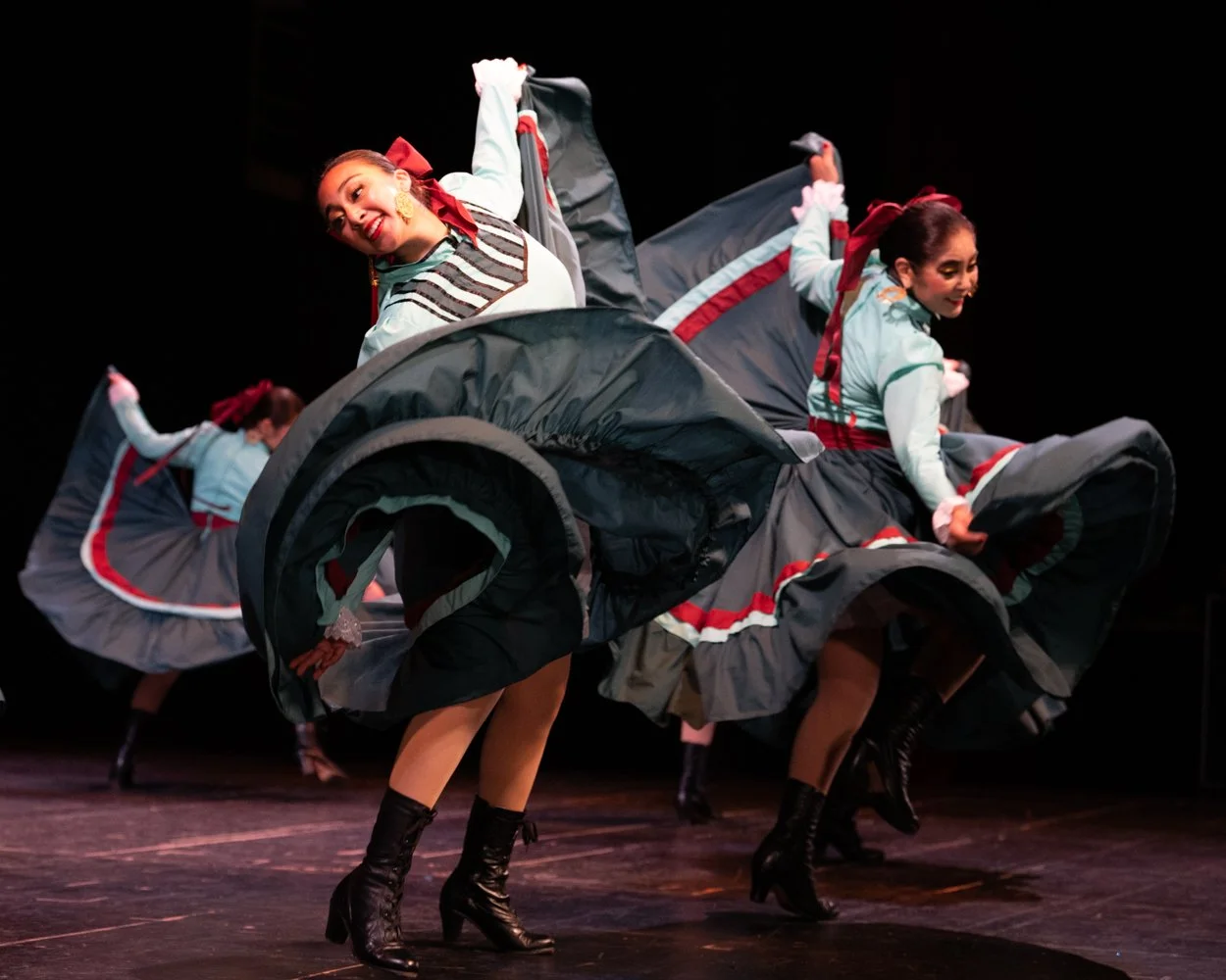 Women performing a traditional dance on stage wearing colorful costumes with full skirts, black boots, and red ribbons in their hair.