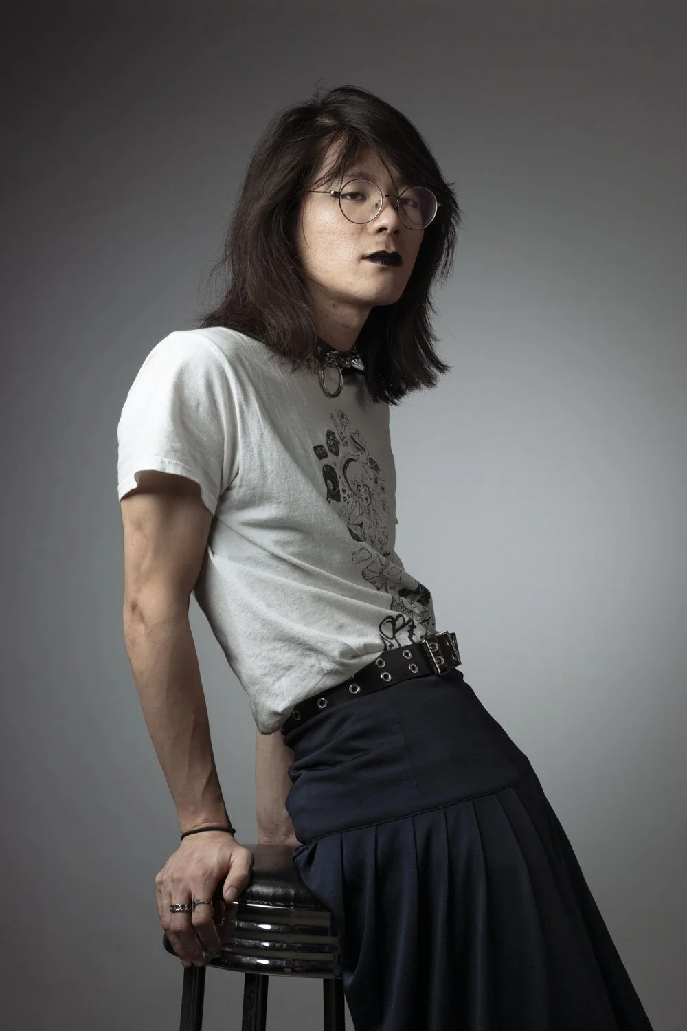 Person with long dark hair, glasses, dark lipstick, wearing white graphic t-shirt and black skirt sitting on a stool against gray background.