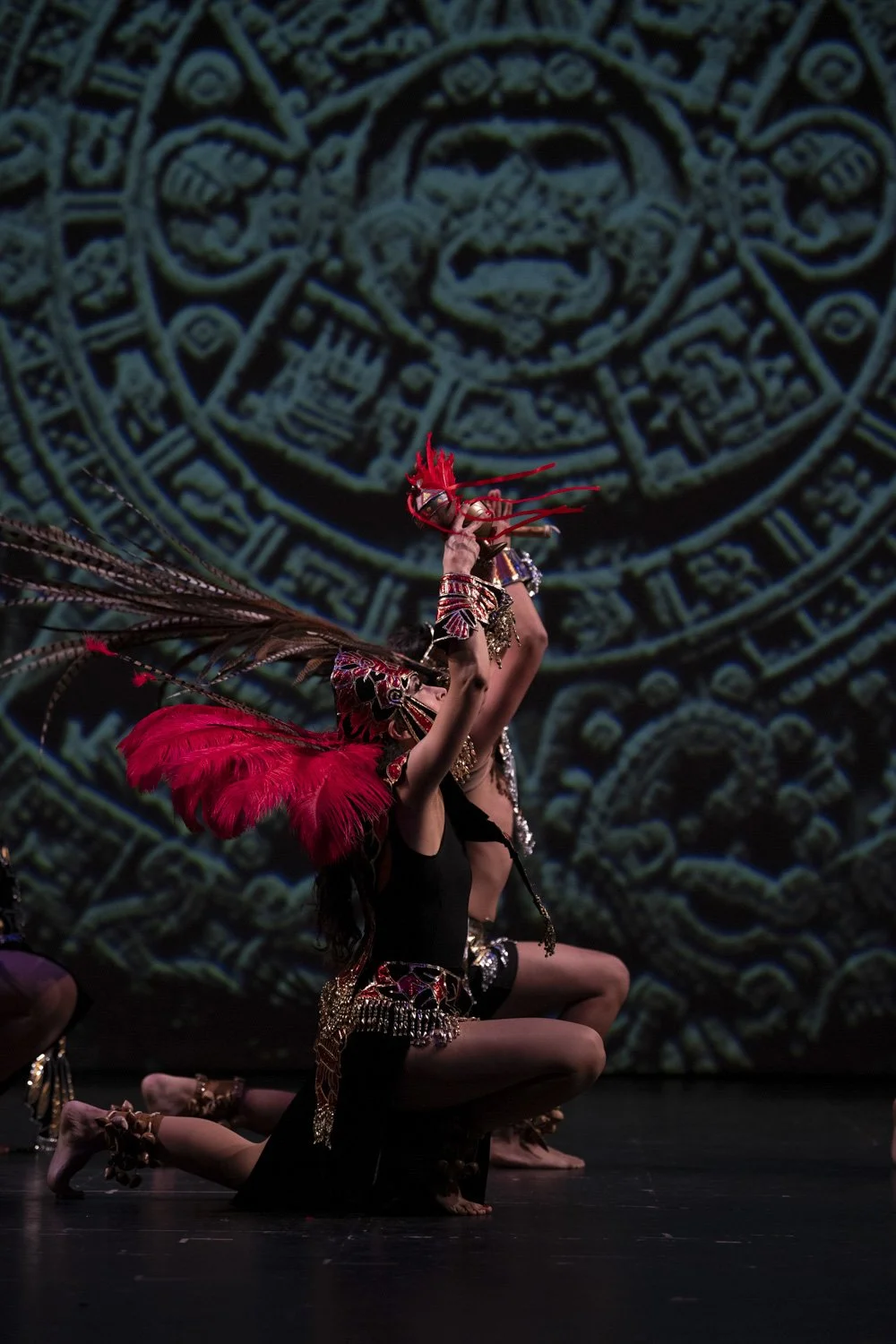 Performer in colorful Indigenous costume with feathers, jewelry, and face paint kneeling on stage during cultural dance with Aztec calendar mural.