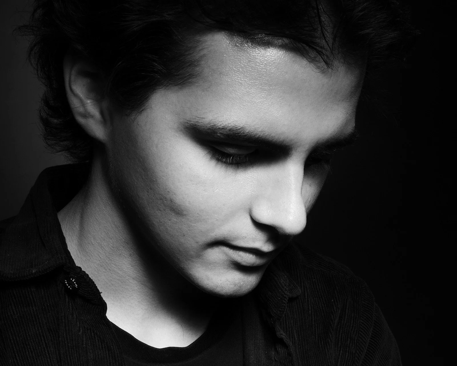 Black and white portrait of young man with closed eyes wearing dark shirt against dark background.