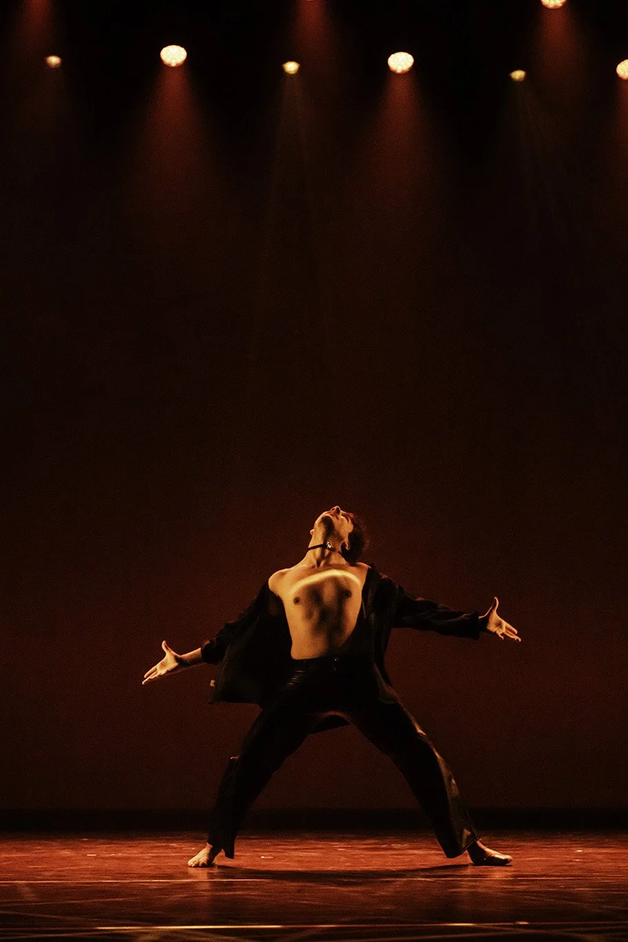 Male dancer performing on stage with dramatic red lighting, shirtless, black pants, arms extended and back arched.