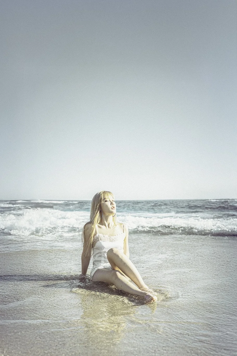 A young woman with long blonde hair sitting on the wet sand at the beach, with her legs crossed and eyes closed, facing the sun, with ocean waves in the background.
