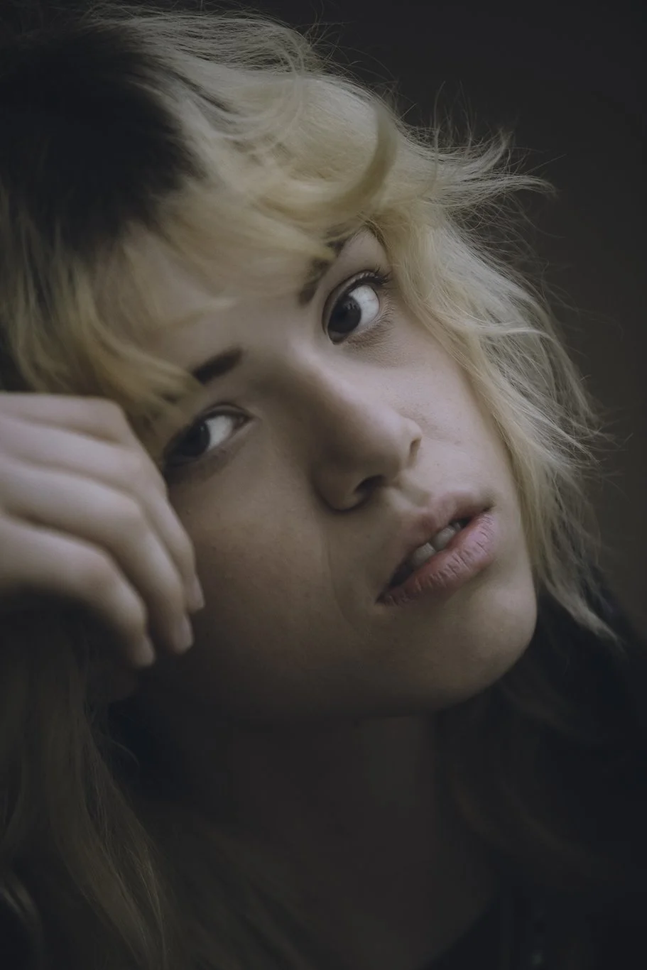 Close-up of young woman with wavy blonde hair looking contemplative, resting head on her hand.