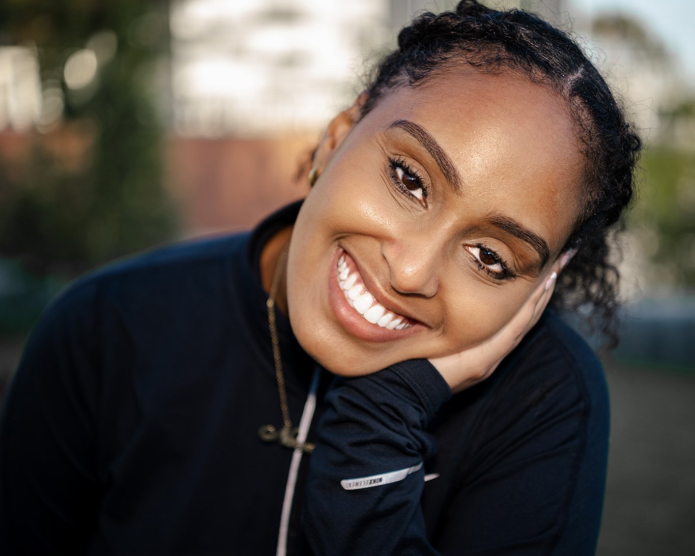 Smiling woman with curly hair in a black jacket resting head on her hand outdoors.
