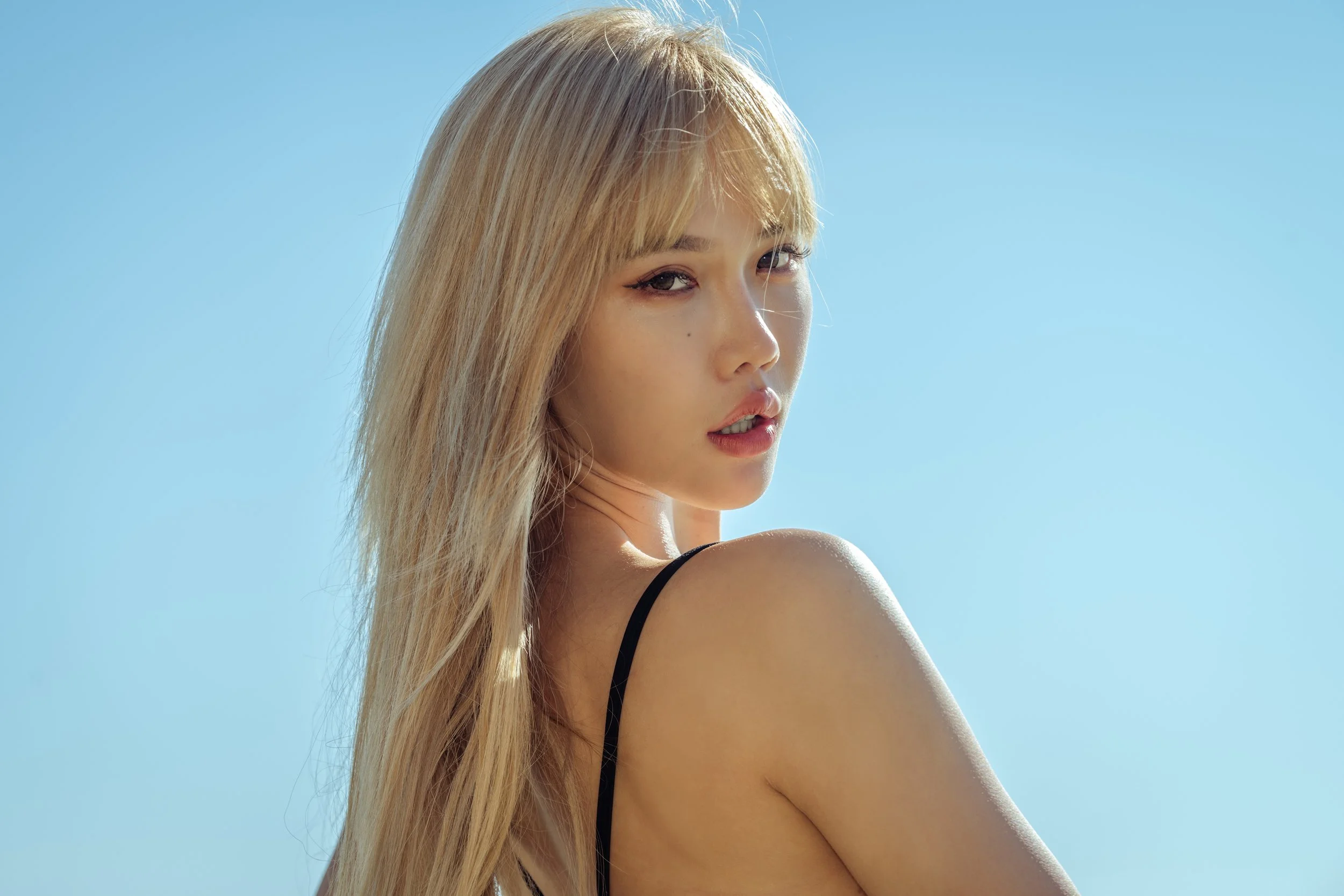 Young woman with long blonde hair wearing a black strap top against a clear blue sky.