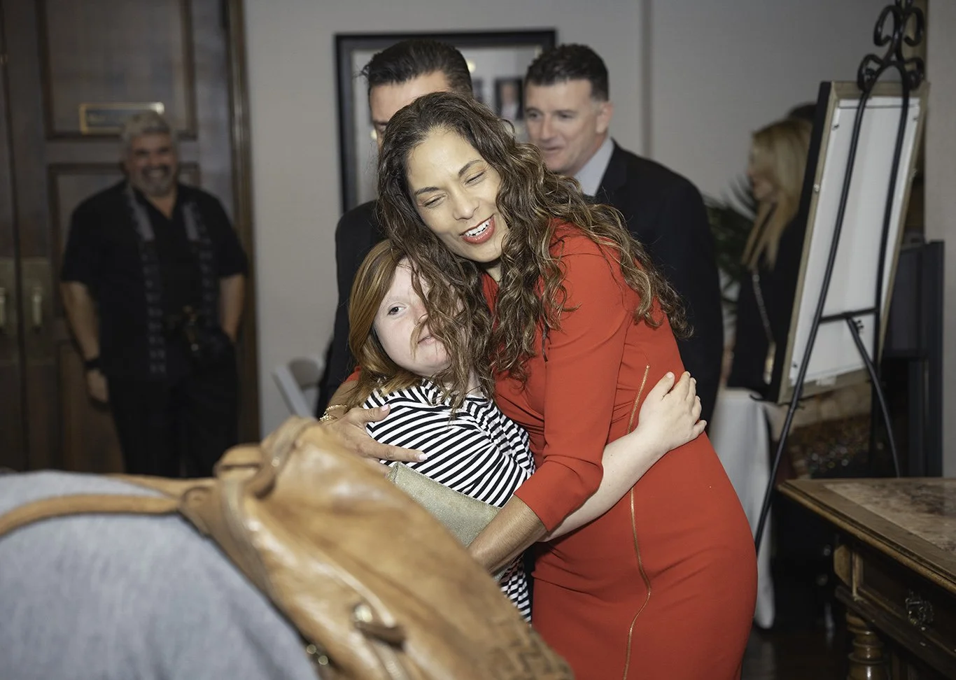Woman in red dress hugging young girl in striped shirt at indoor gathering.