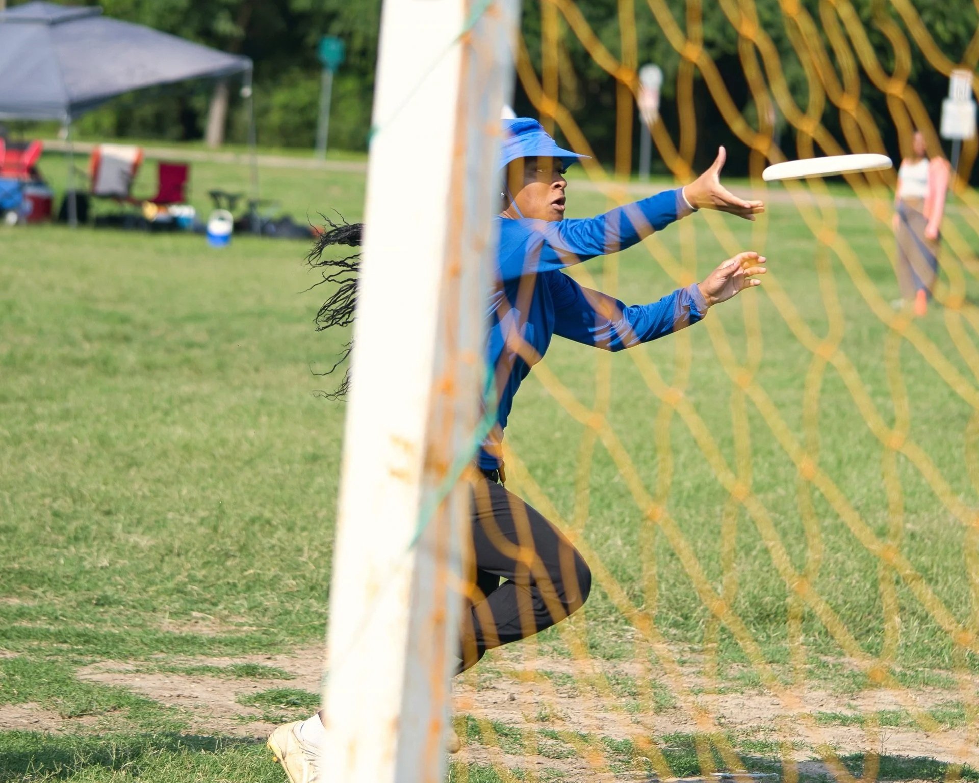 A woman in blue athletic clothing and a hat is playing disc golf, catching a white disc near the goal net.