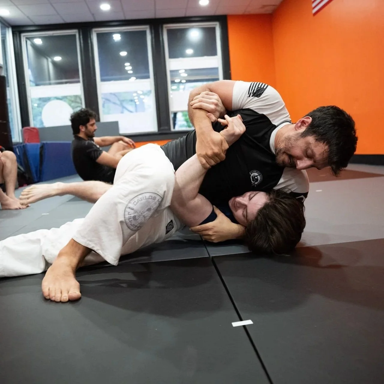 Two students of Brooklyn Heights Jiu Jitsu sparring together during class, with other students sitting on the mats behind them.