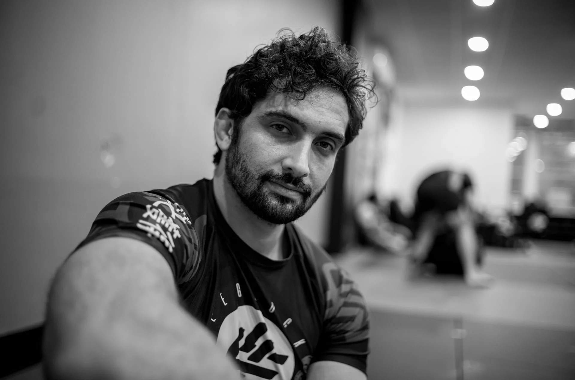 A picture of a Jiu Jitsu practitioner smiling, with students training Jiu Jitsu behind him.