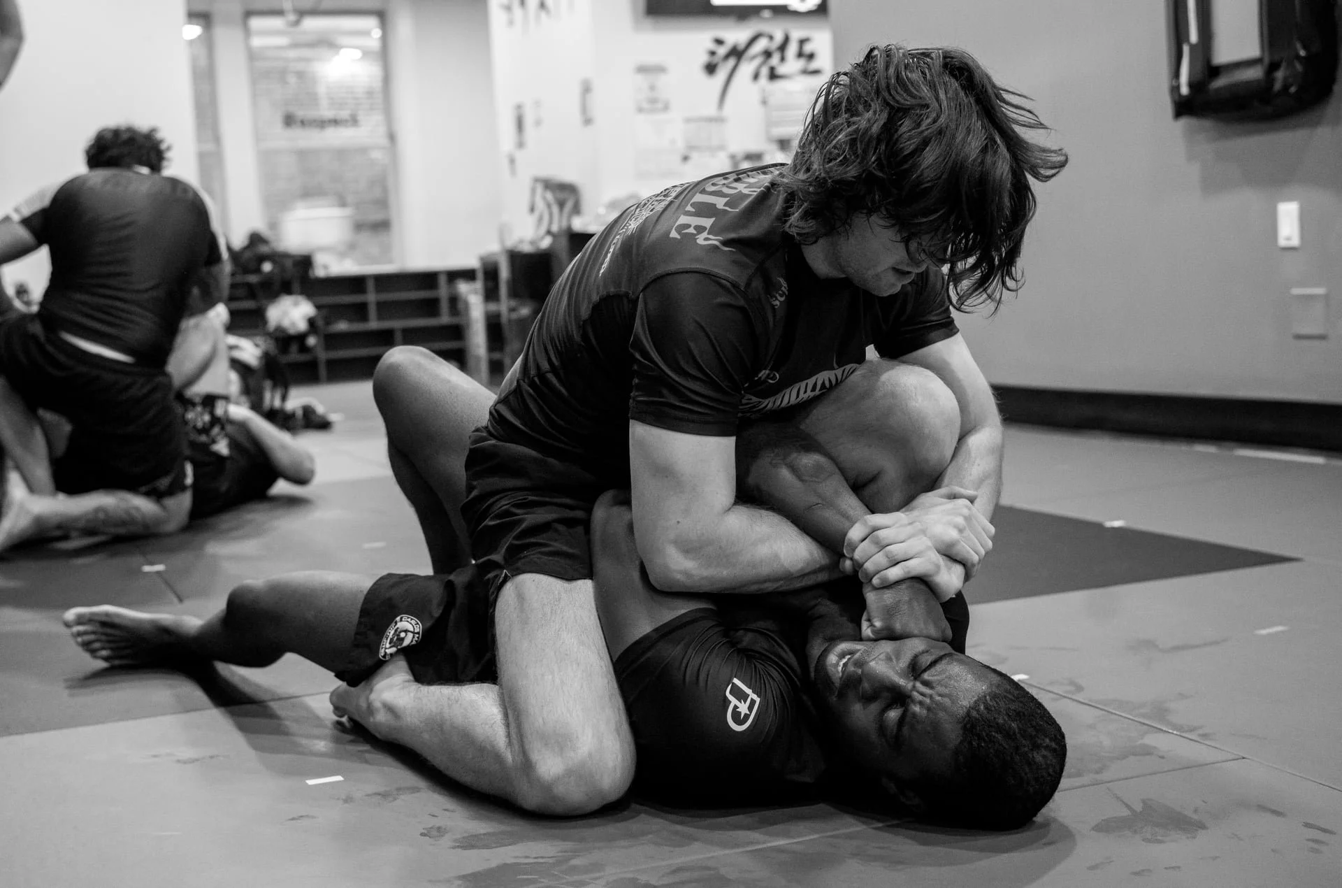 Two pairs of Jiu Jitsu students grappling on the mat during a training session in a gym, with one pair in the foreground and the other in the background.