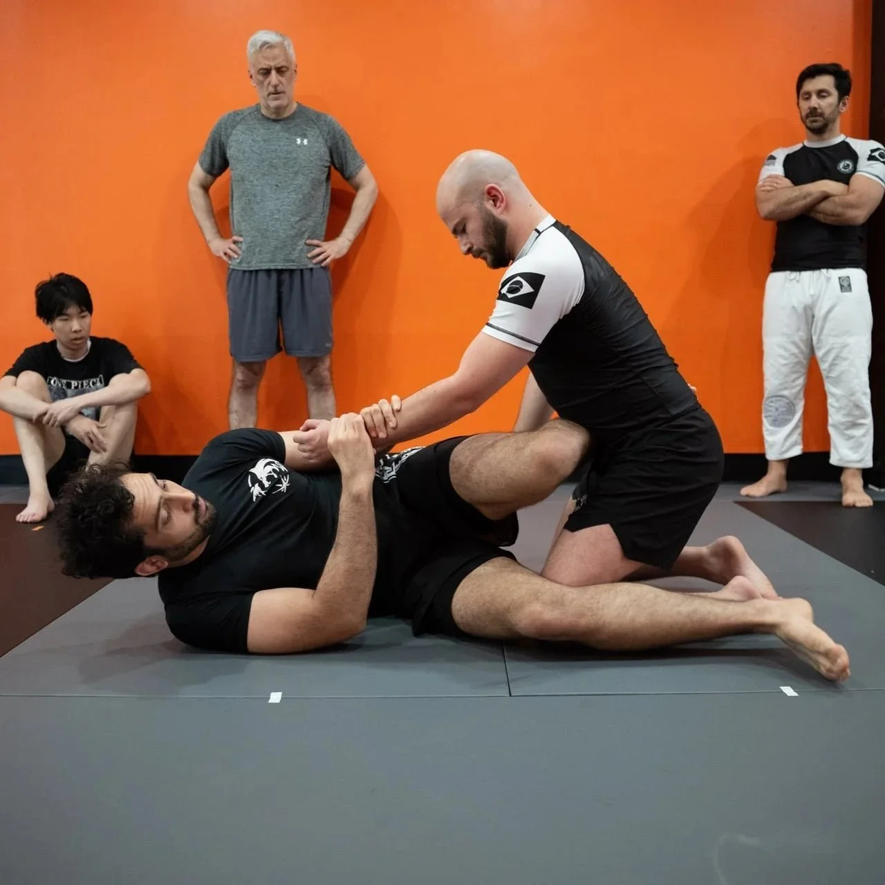 Two men practicing Brazilian Jiu-Jitsu during a jiu jitsu class, with the instructor demonstratng a jiu jitsu technique. Three students watch.