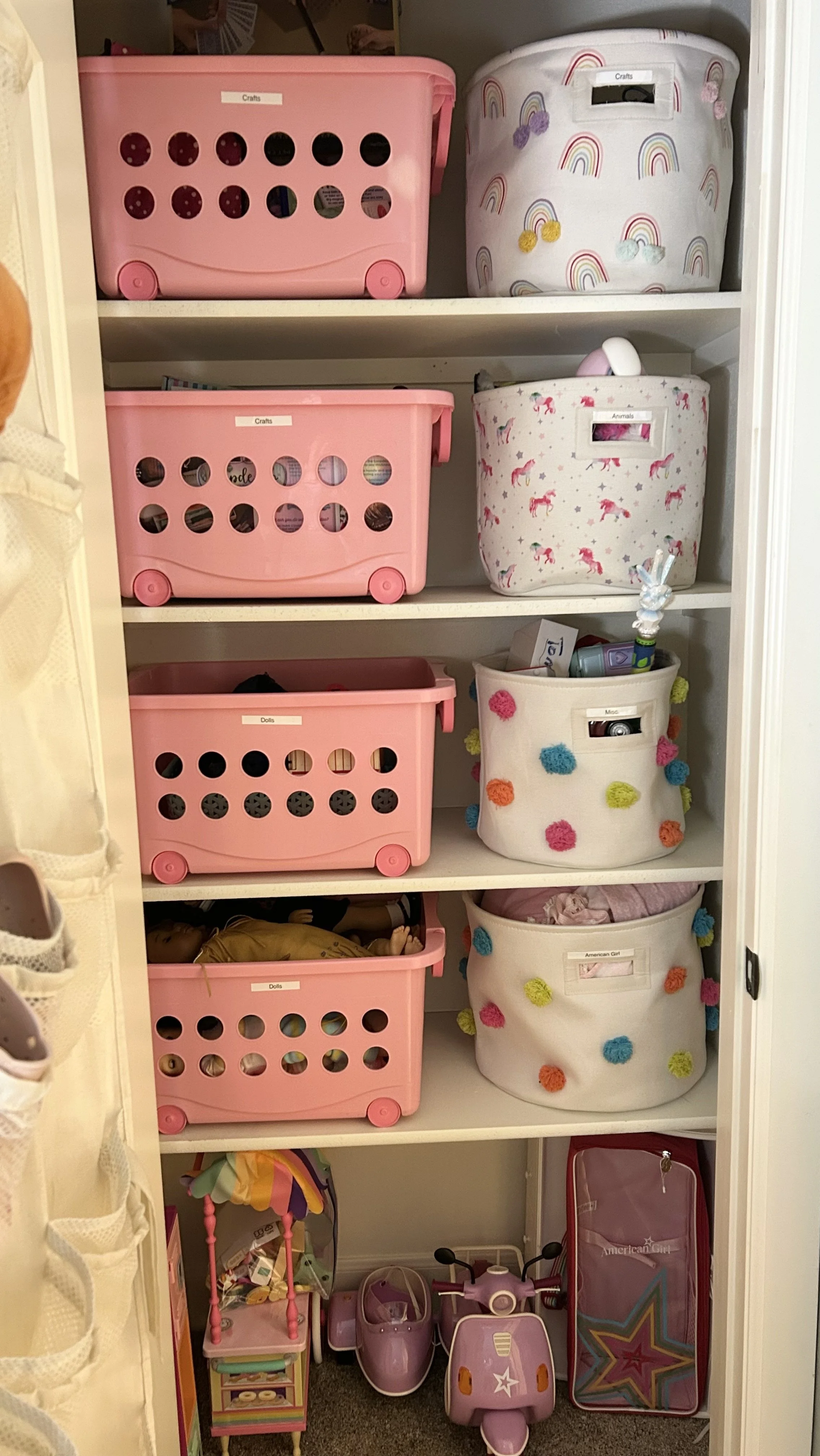 Closet with four shelves containing toys and storage bins. The top three shelves have pink plastic bins labeled for crafts, animals, dolls, and miscellaneous items. The bottom shelf holds a dollhouse, a toy scooter, a ride-on vehicle, and an American