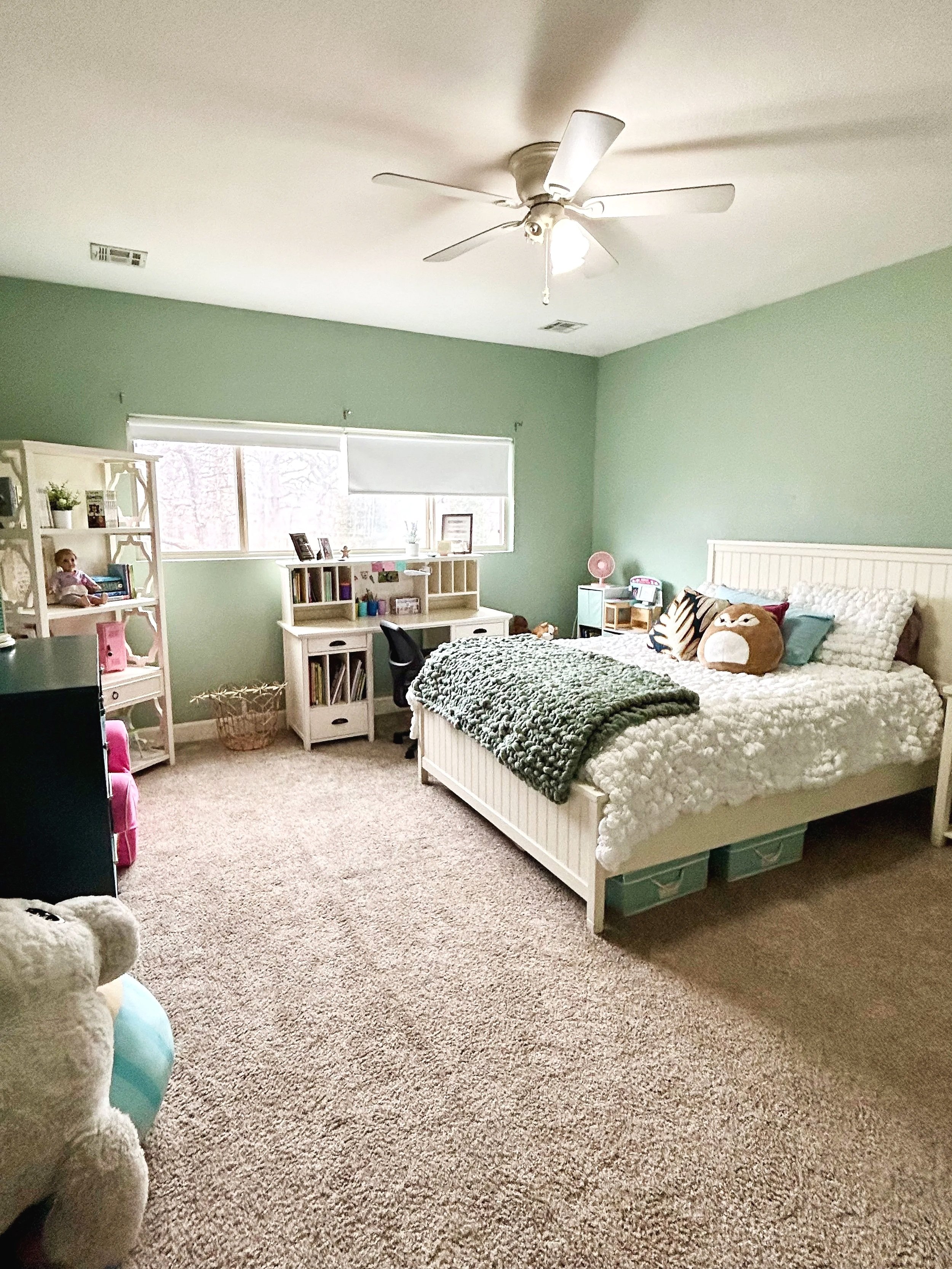 A bedroom with a white bed, green walls, a ceiling fan, a desk with a black chair, shelves with books and decorations, and plush toys on the bed.