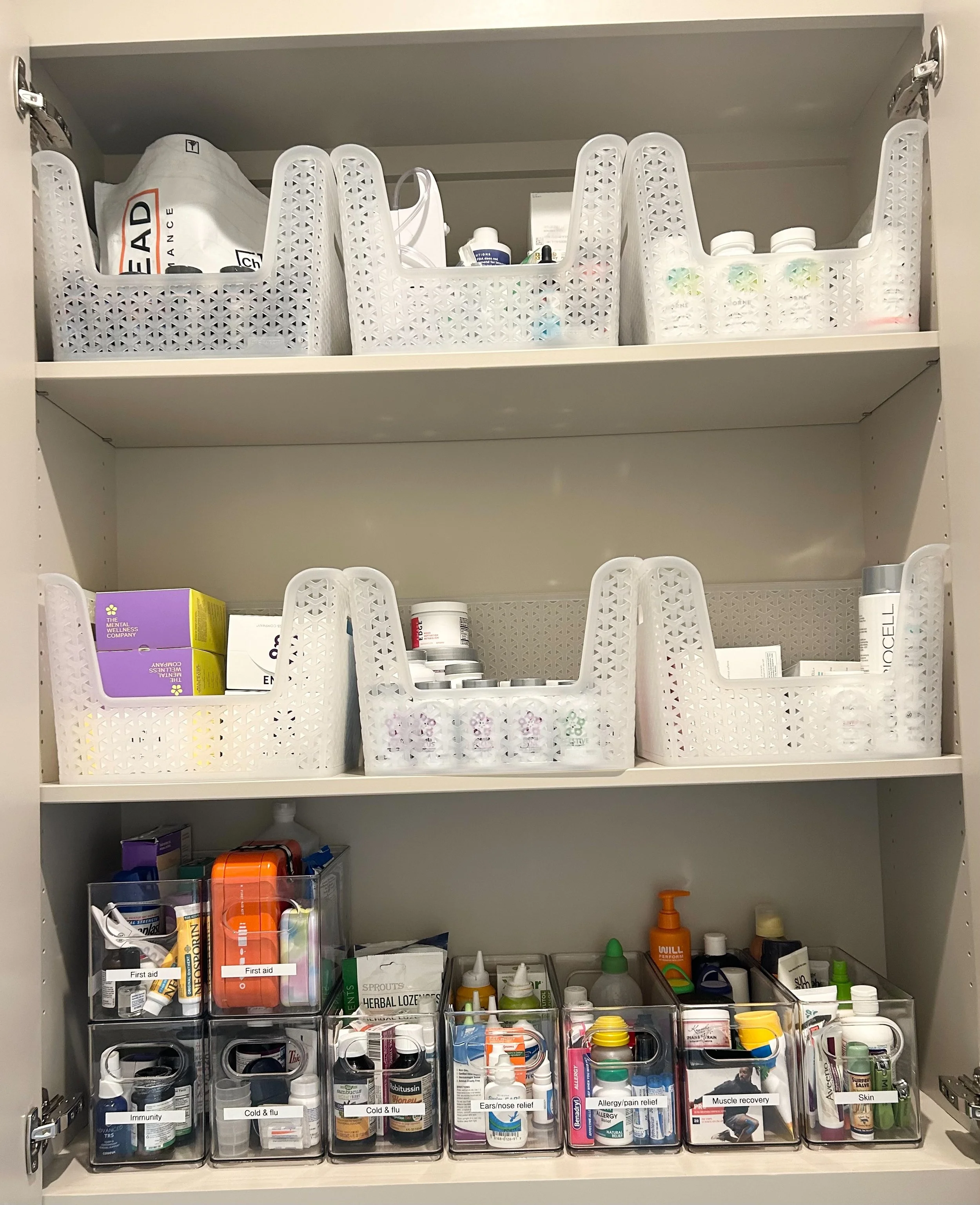 Organized medicine cabinet with white plastic baskets holding various medications and supplies on three shelves, and clear containers with labeled medications for first aid, cold and flu, allergy and pain relief, muscle recovery, and skin care on the