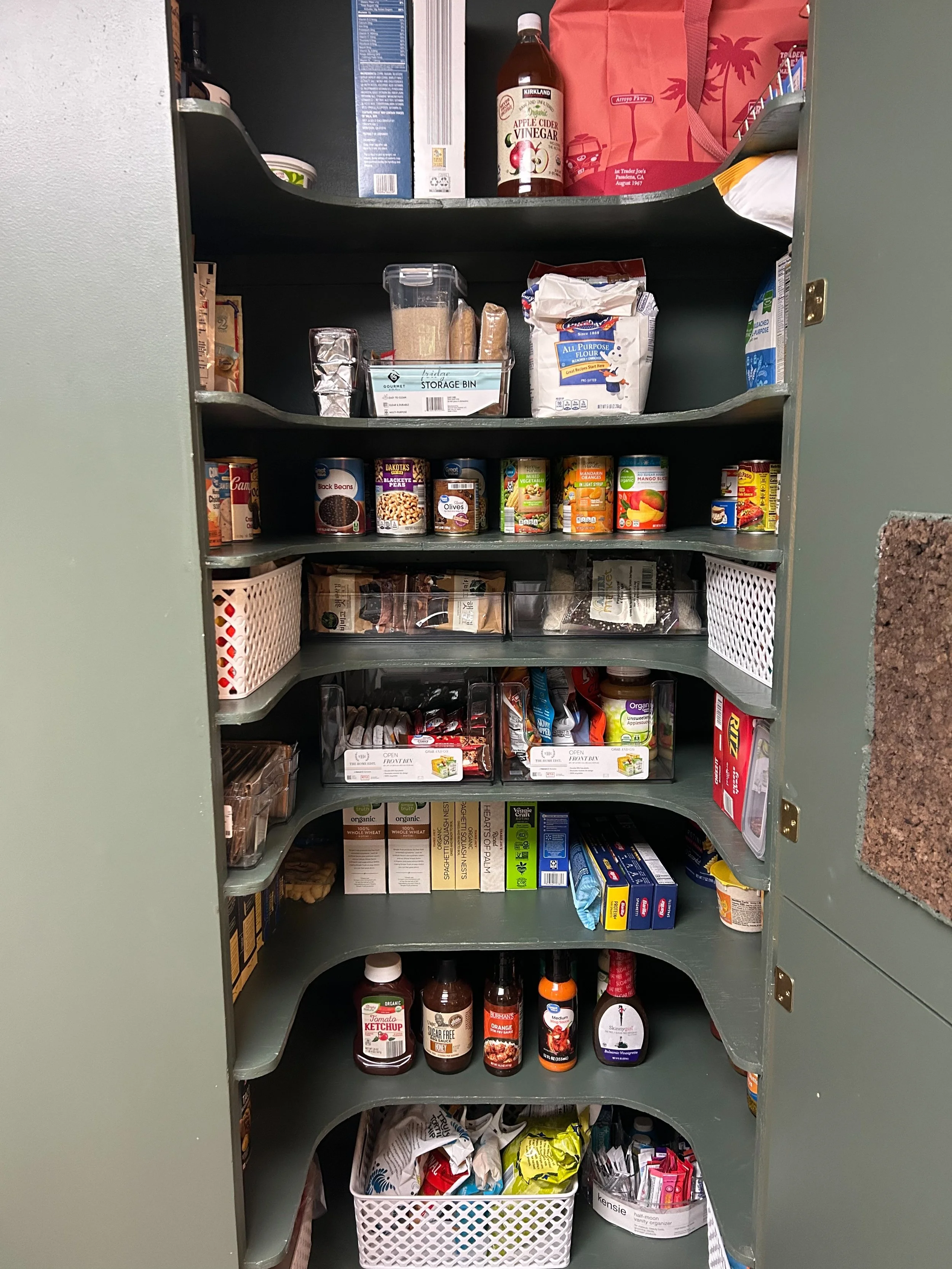Open pantry with various canned goods, boxes, bottles, and snack items stored on multiple shelves and in baskets.