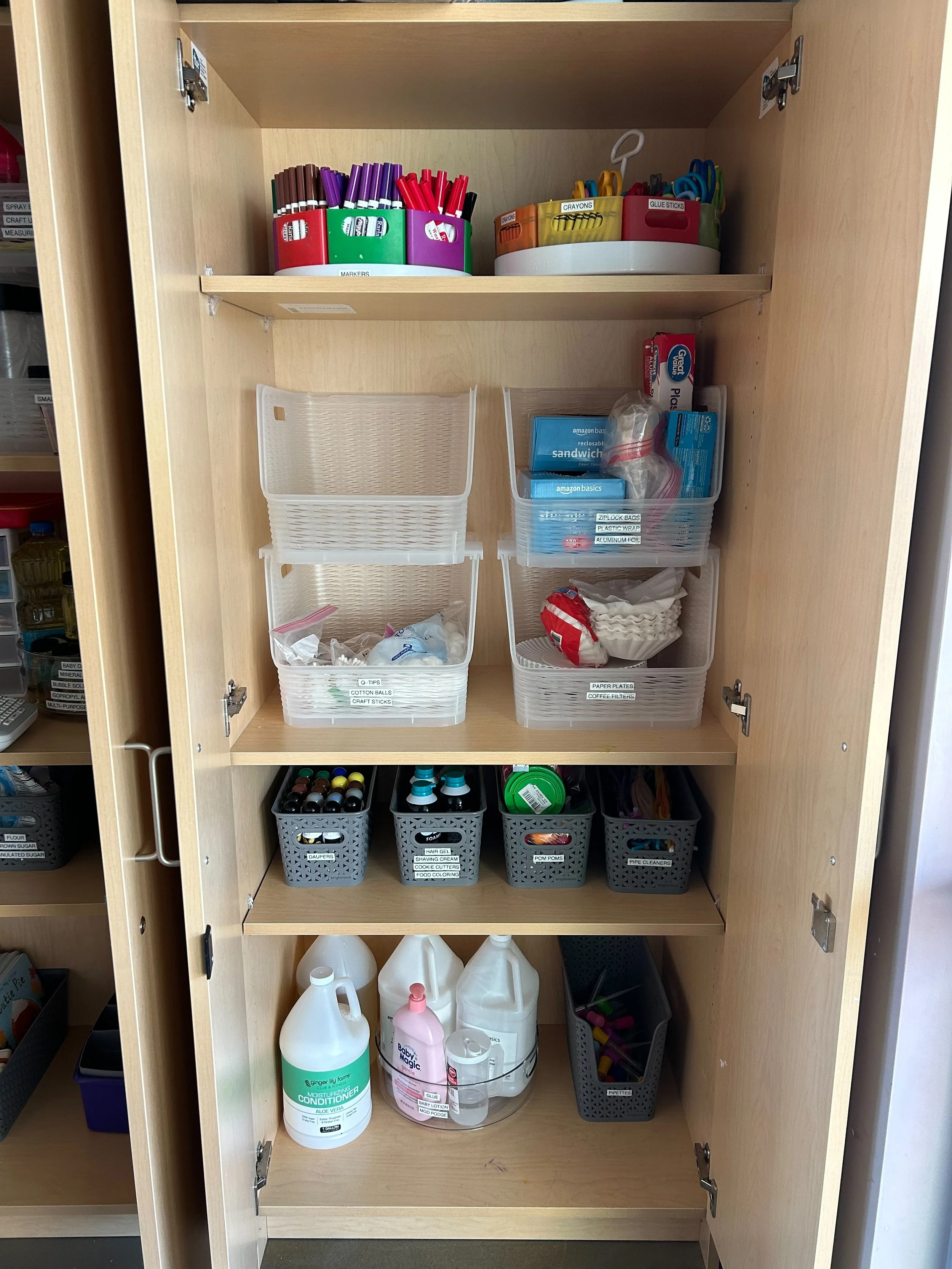 A wooden cabinet with four shelves containing various craft and office supplies, including markers, scissors, glue sticks, plastic bags, cotton balls, paper plates, cleaning supplies, and small containers with different items.