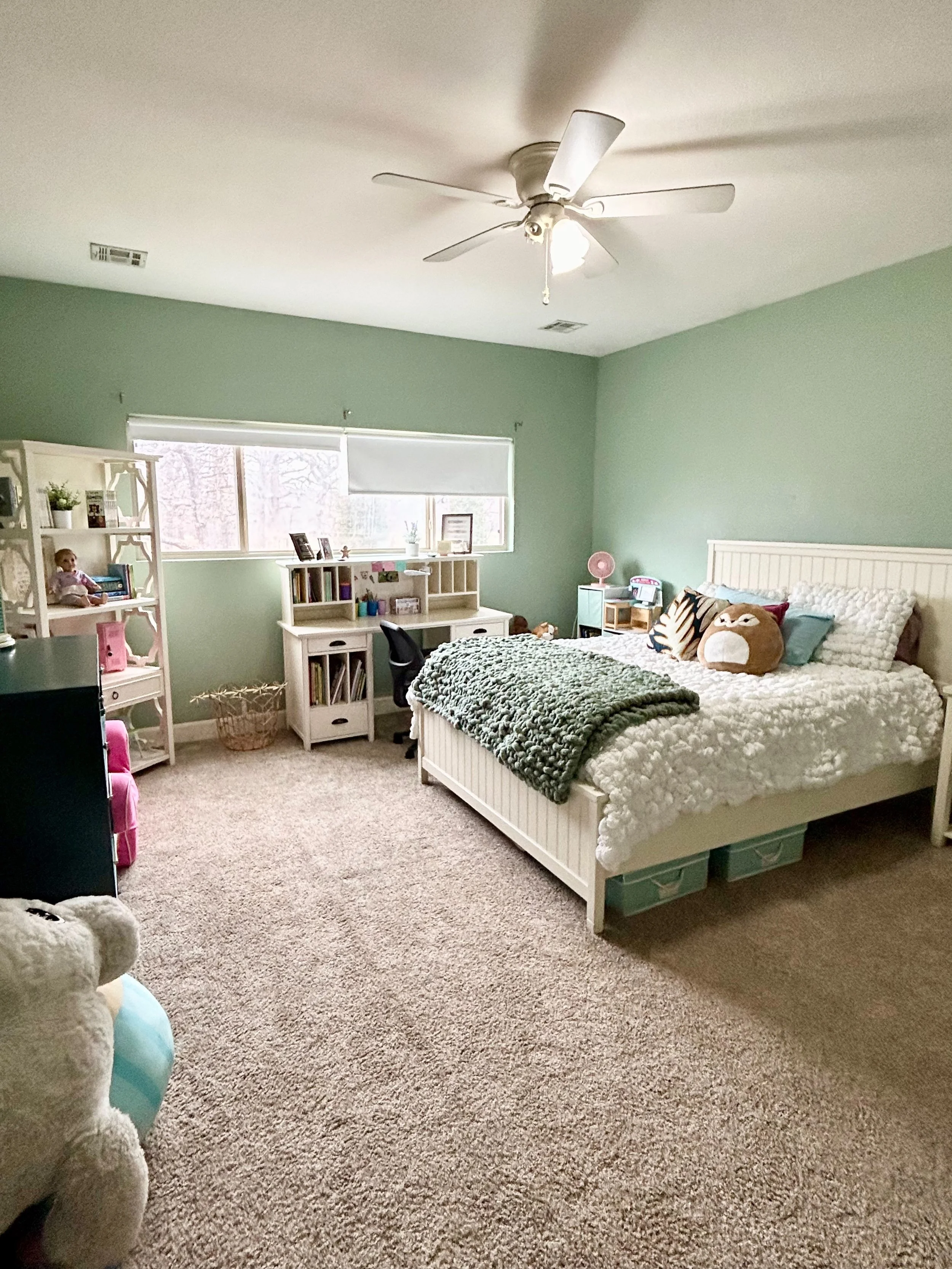 A bedroom with a white bed, plush pillows, and a textured blanket. A ceiling fan with lights, large windows, a desk with a chair, bookshelves, and various toys and decor.