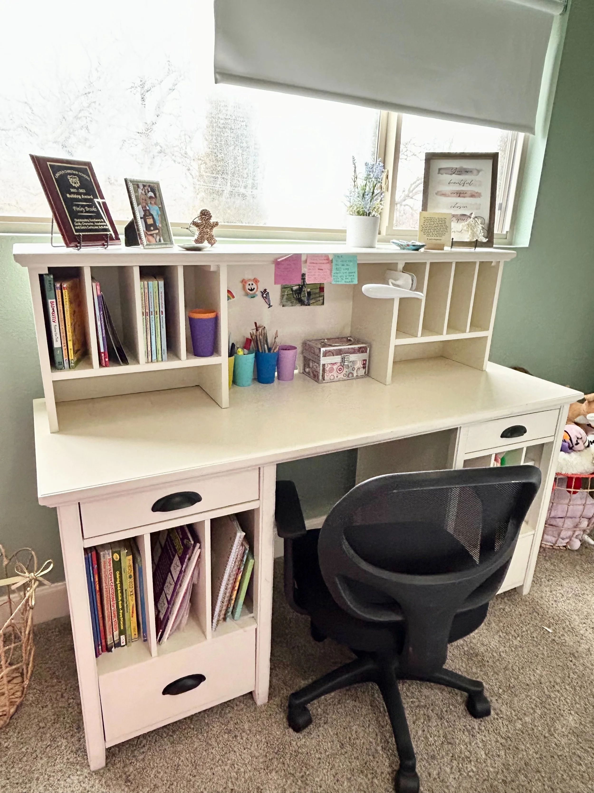 A white desk with multiple storage compartments and a black office chair in front. The desk has a higher shelf with picture frames, books, and decorative items, and a window behind it with a partly rolled-up blind. The bookshelf on the left contains 