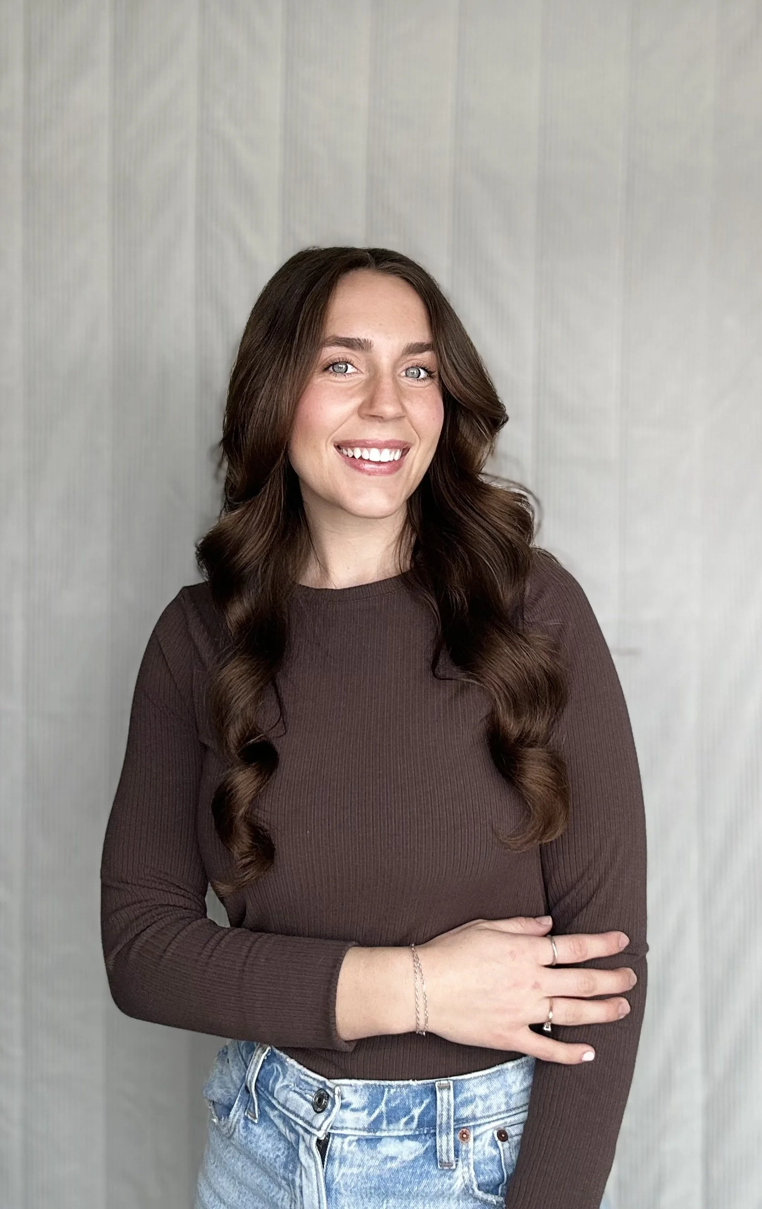 A smiling woman with long, wavy brown hair wearing a brown long sleeve shirt and blue jeans, standing against a light-colored textured background.
