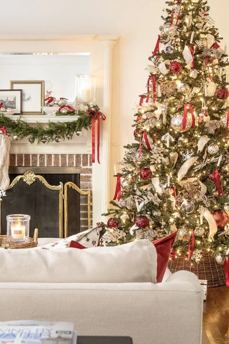 Decorated Christmas tree with red, gold, and silver ornaments, ribbons, and lights in a cozy living room with a brick fireplace, a mantel with greenery and candles, and a sofa with red and white pillows.