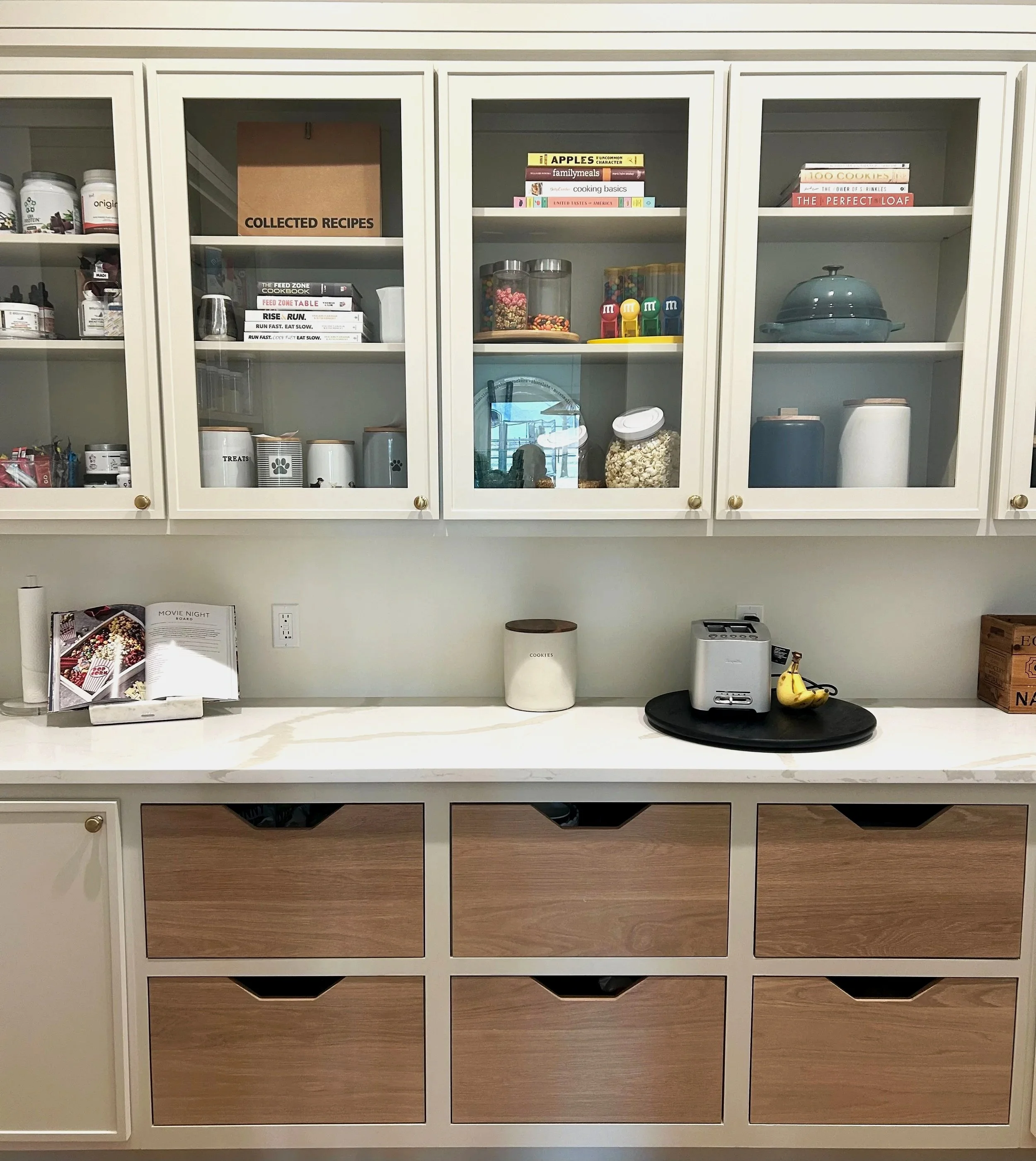 Kitchen counter with wooden drawers, a white countertop, a microwave, bananas, a canister, and a cookbook, with white cabinets above containing jars, books, and containers.