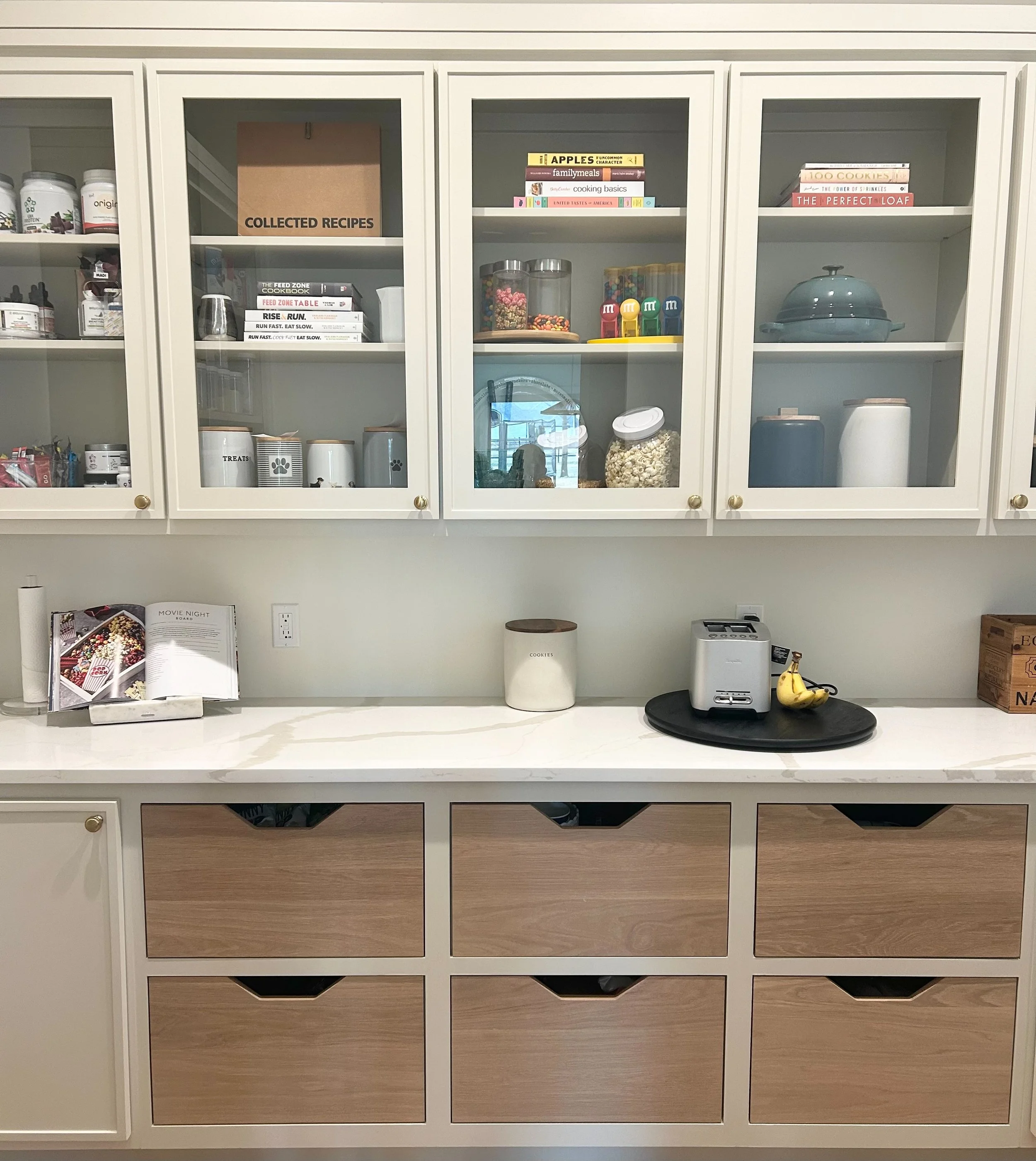 Kitchen counter with open white cabinets above containing jars, books, and snack items; countertop with cookbooks, cookie jar, toaster, bananas, and a tray; drawers below.