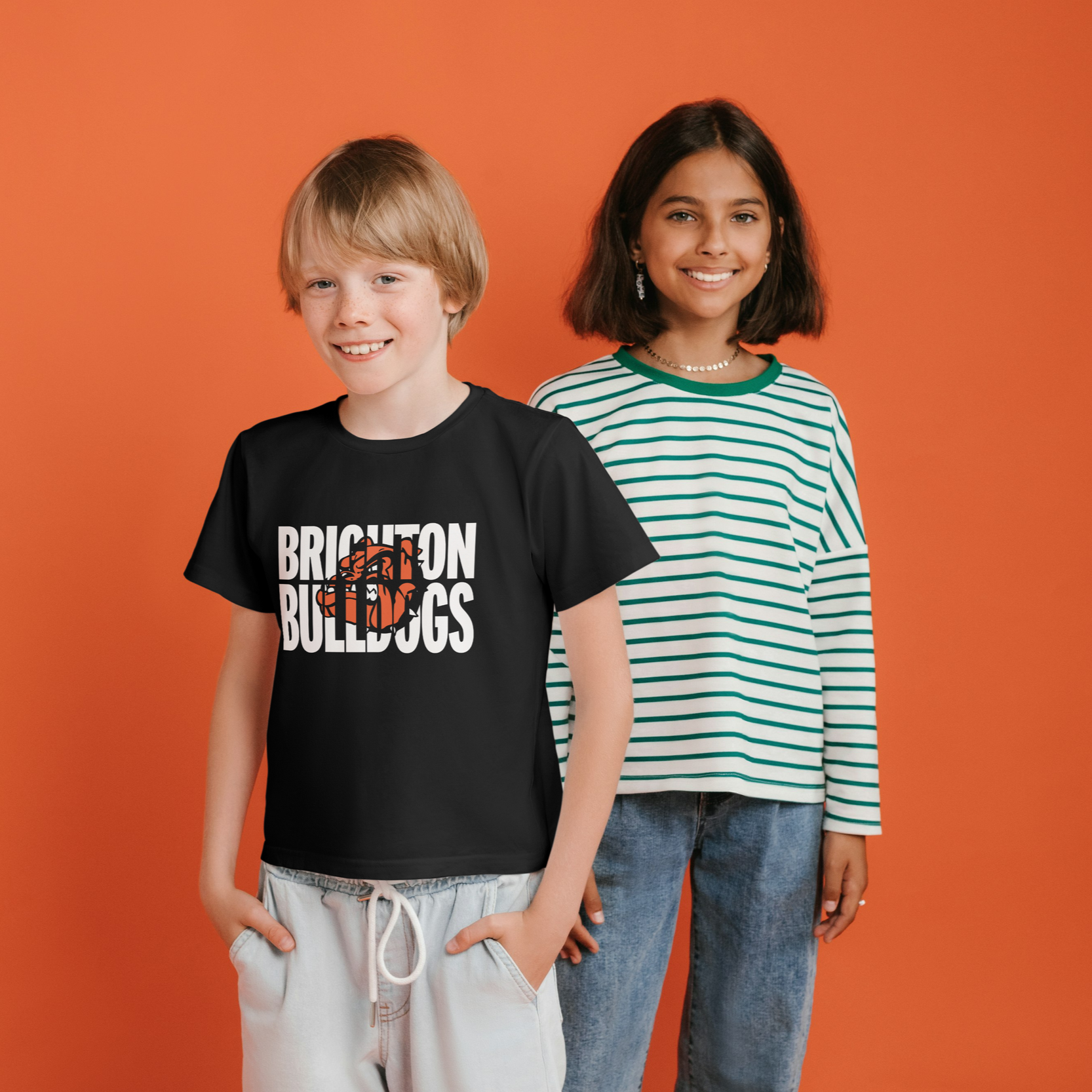 A boy and a girl standing in front of an orange background, smiling at the camera. The boy wears a black T-shirt with 'Brighton Bulldogs' logo and light-colored shorts. The girl wears a striped green and white long-sleeve shirt and blue jeans.