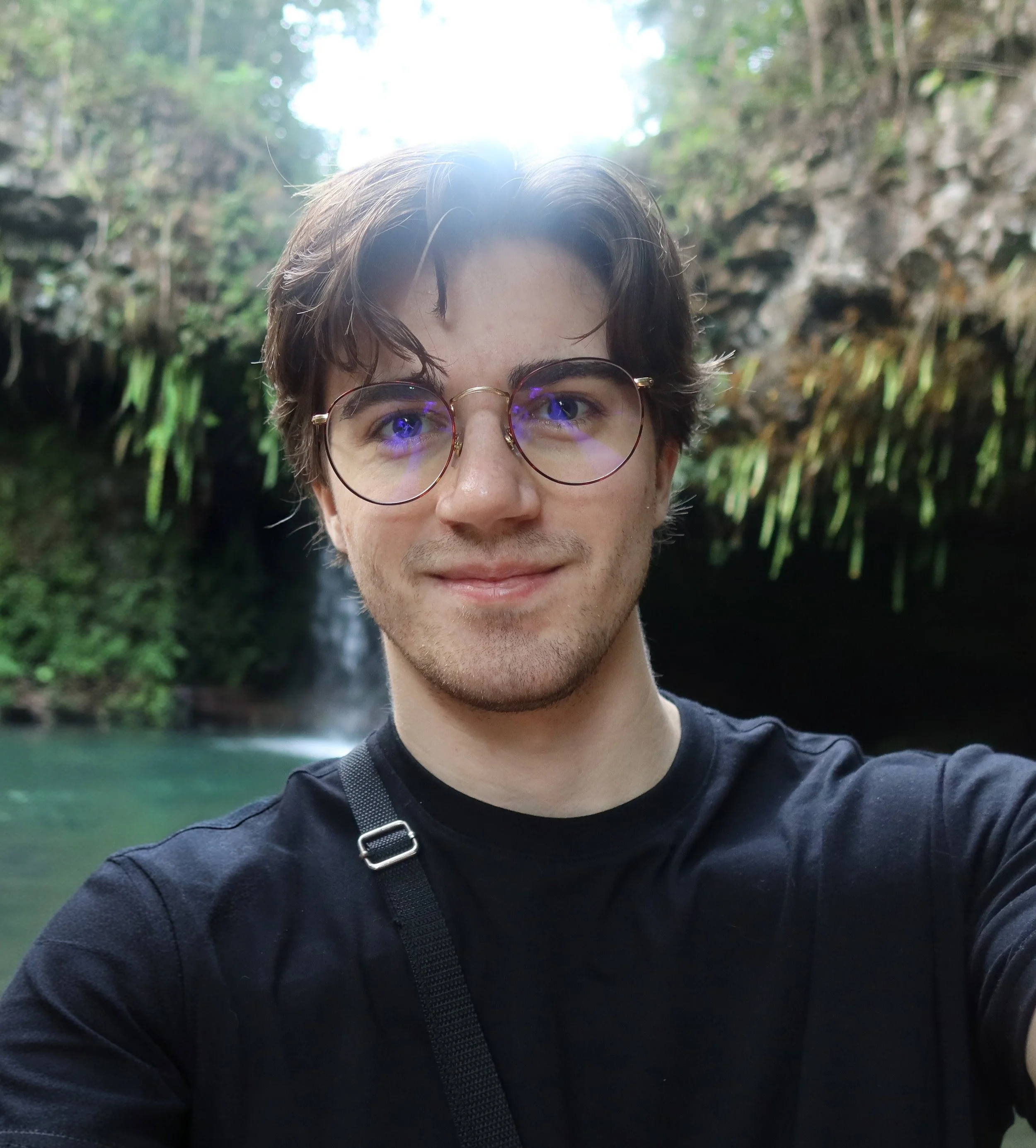 A young man with glasses taking a selfie outdoors in front of a waterfall and greenery.