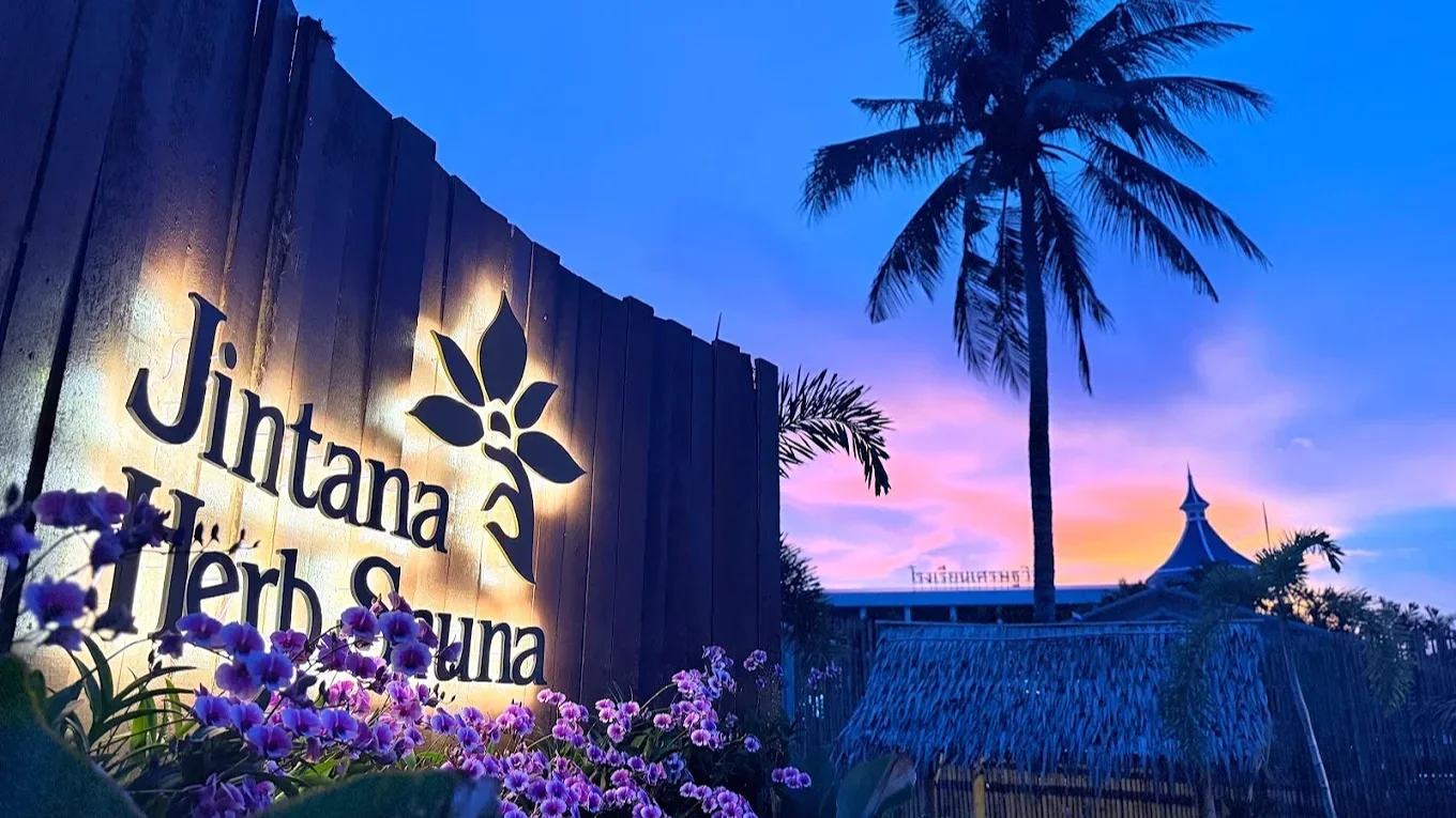 Sunset sky with pink and purple hues, a tall palm tree, a wooden fence with illuminated sign that reads "Jintana Herbal & Therapy," purple flowers in foreground, and a building with a pointed roof in the background.