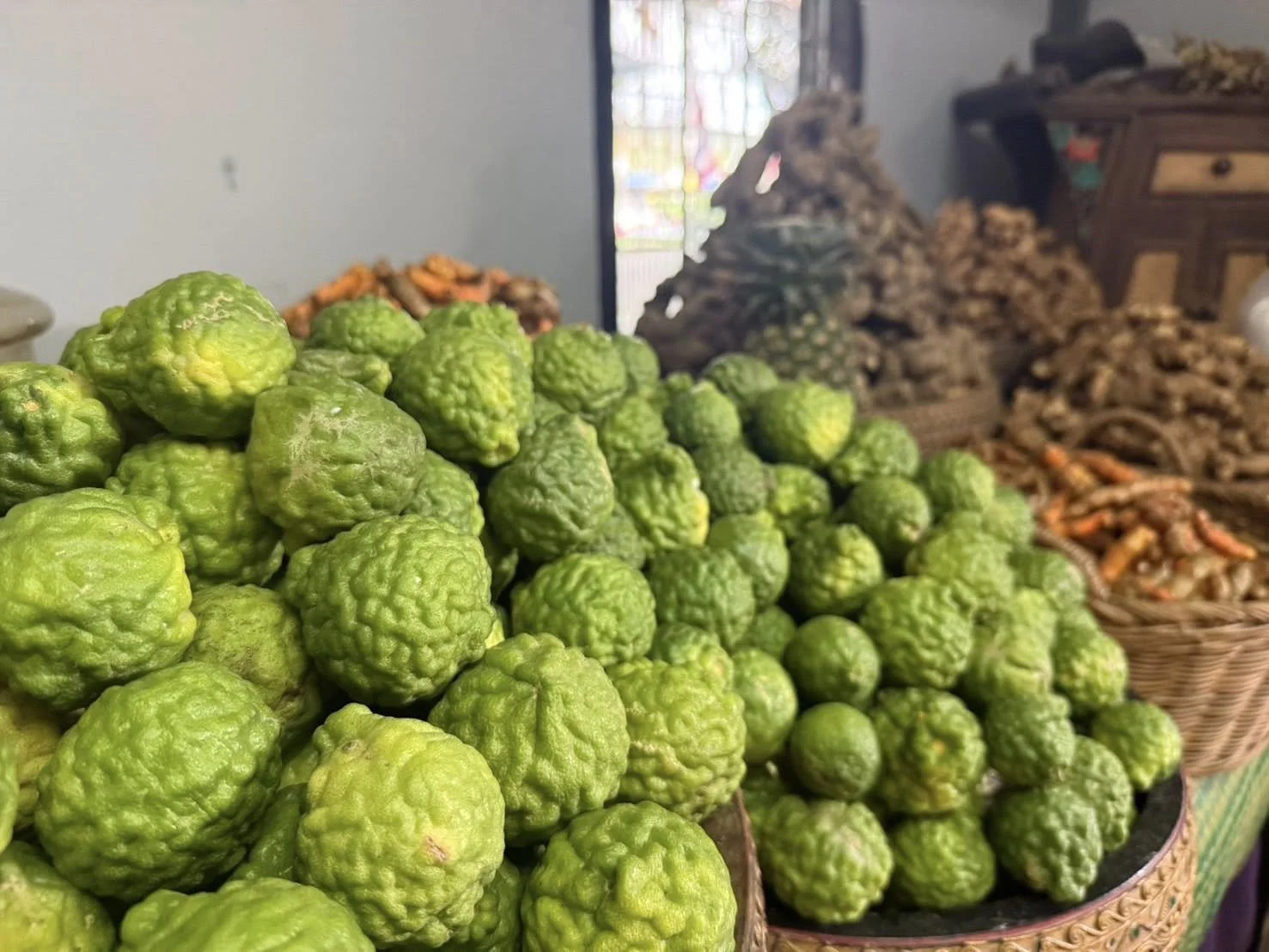 A close-up of green kaffir limes in baskets, with dried ginger and other produce in the background.