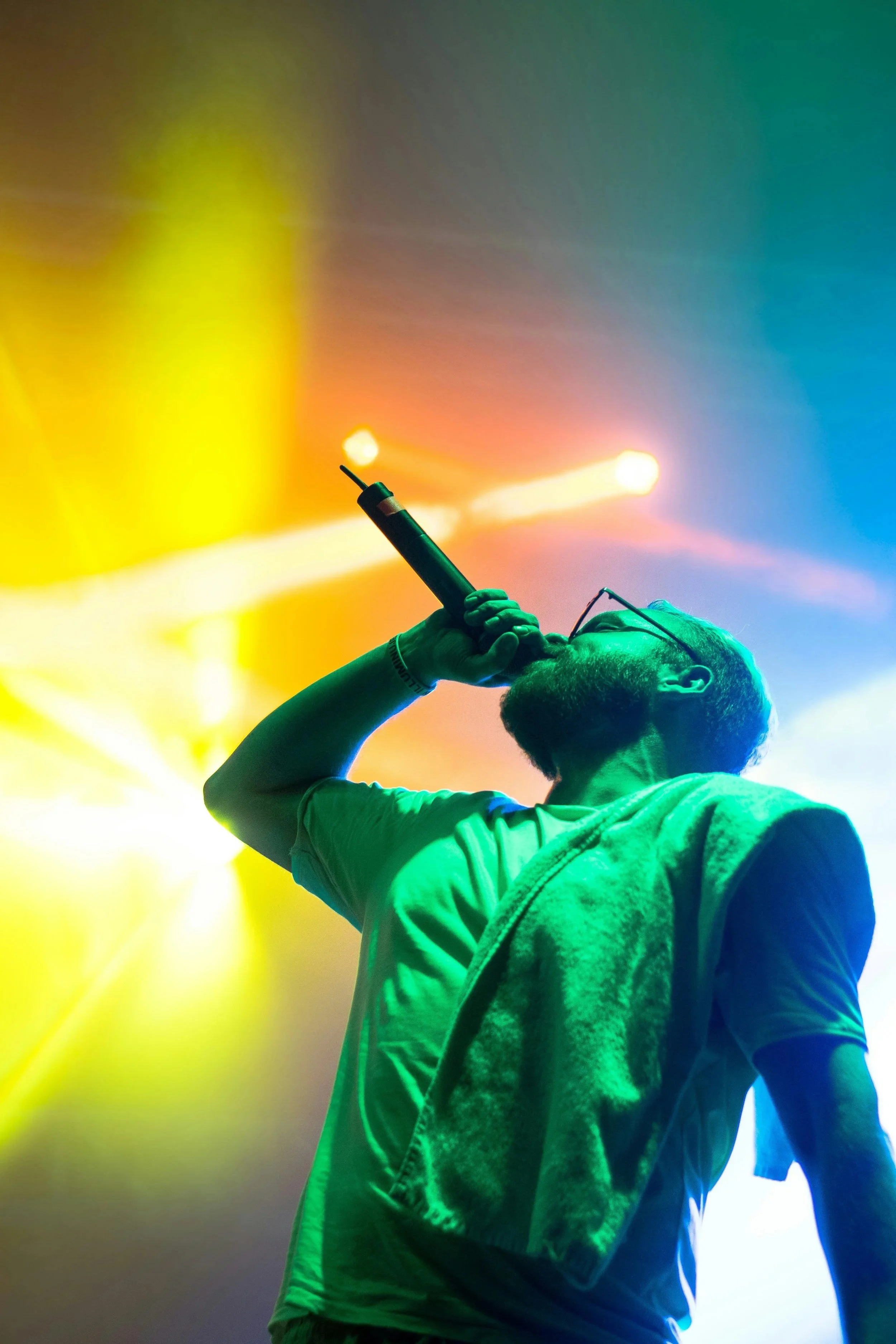 A man with glasses, a beard, and a towel over his shoulder singing into a microphone on stage with colorful stage lighting.