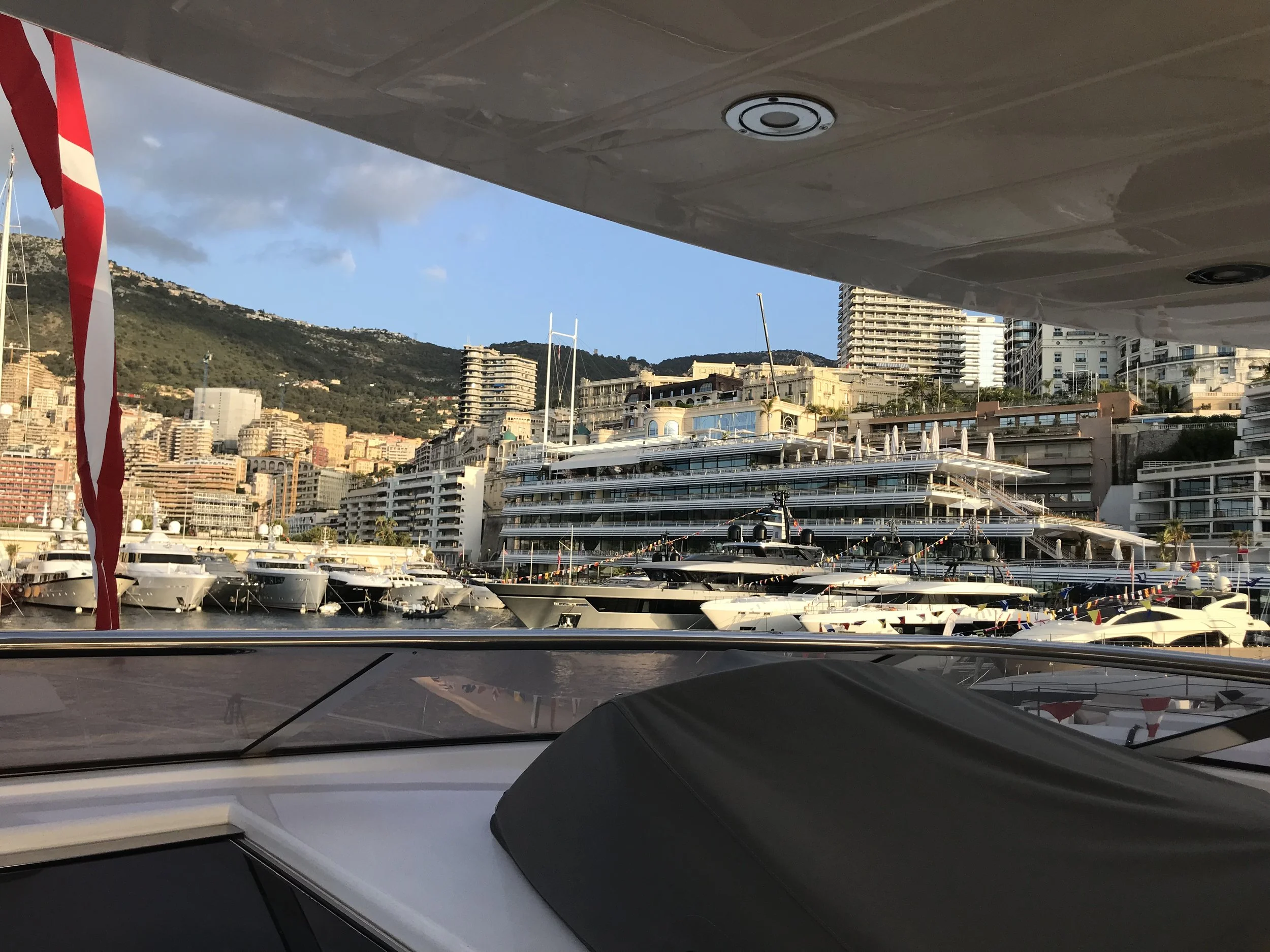 View from inside a yacht looking out over a marina filled with luxury boats and yachts, with a cityscape and hillside in the background during daytime.