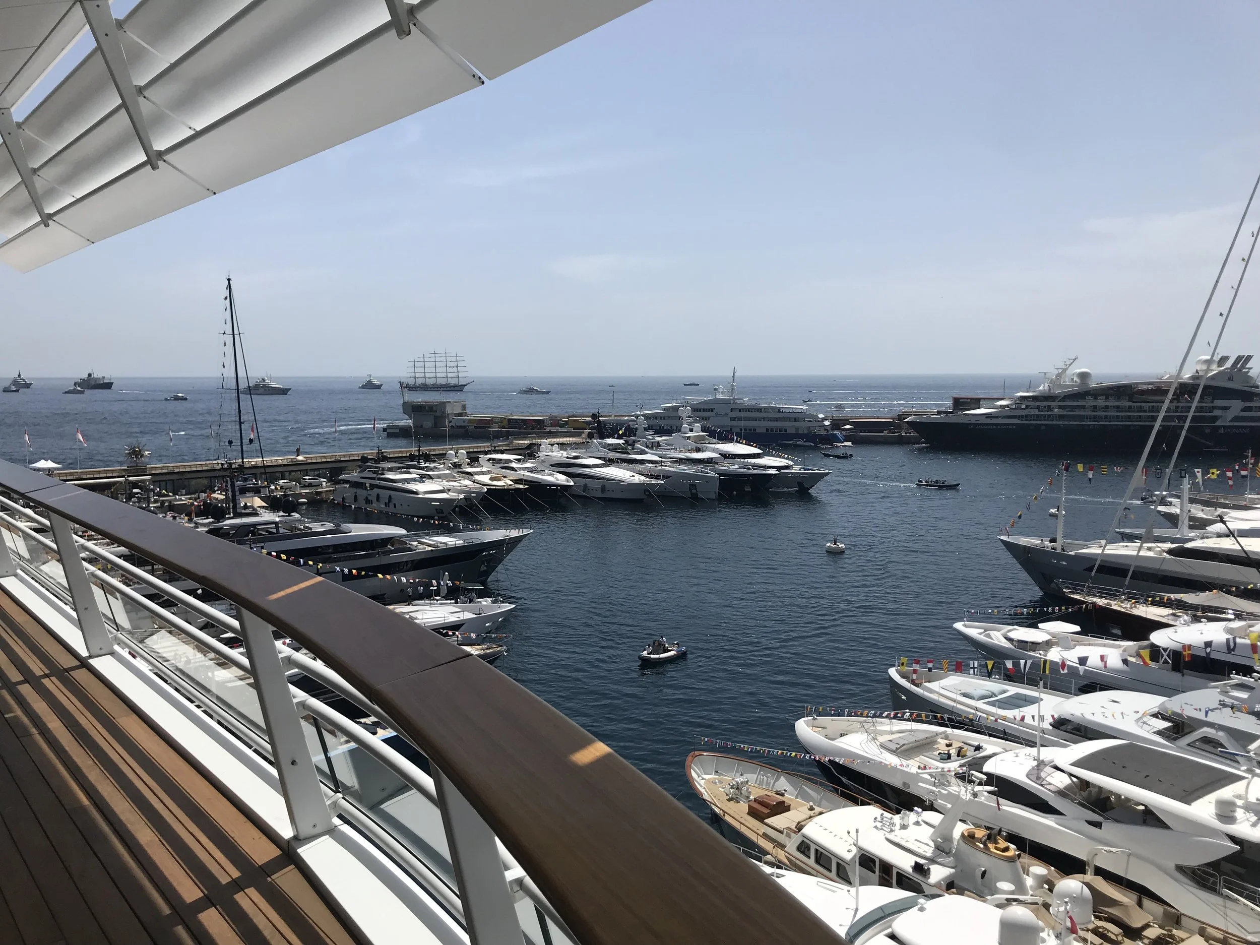 View from a ship's deck overlooking a marina filled with yachts and boats, with the open ocean and a clear sky in the background.