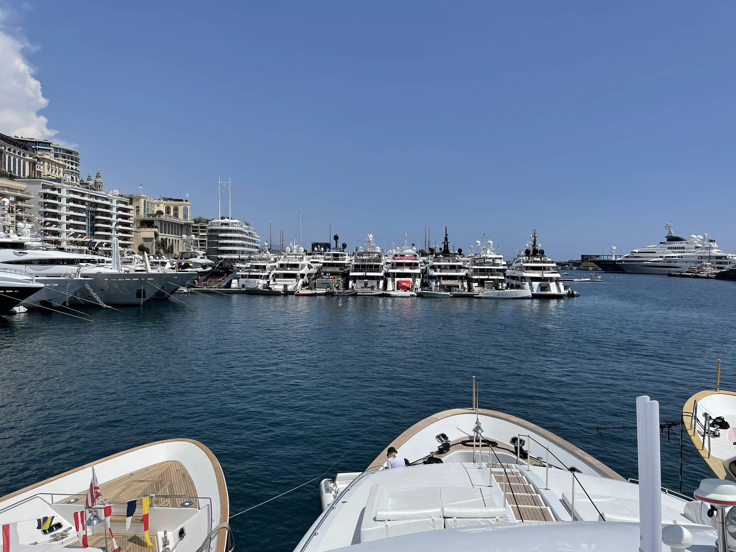 A marina filled with various sizes of luxury yachts docked along a waterfront, with multi-story buildings lining the shore and a clear blue sky overhead.