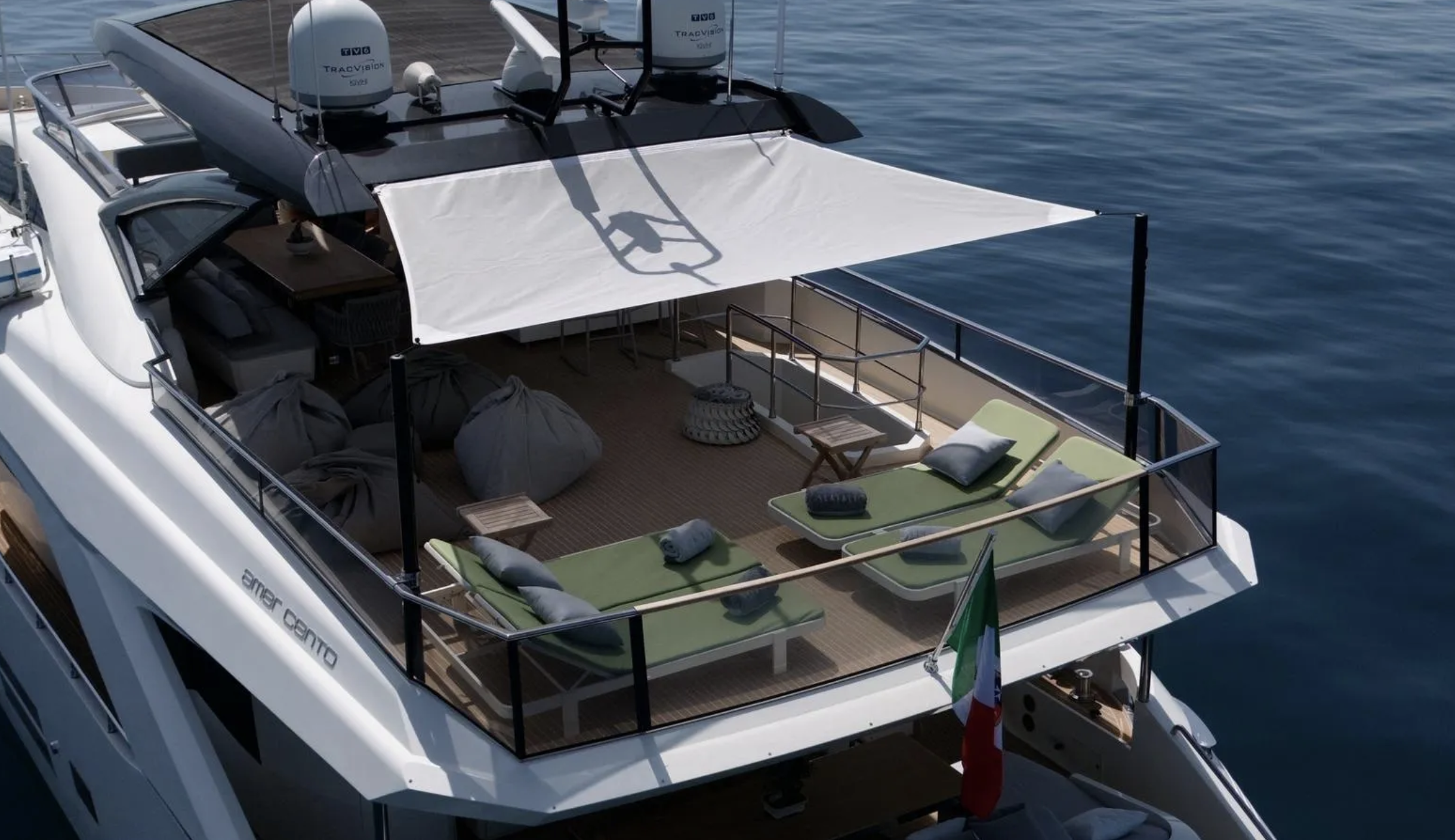The deck of a white yacht with green lounge chairs, pillows, and a shade cover, on calm blue water.