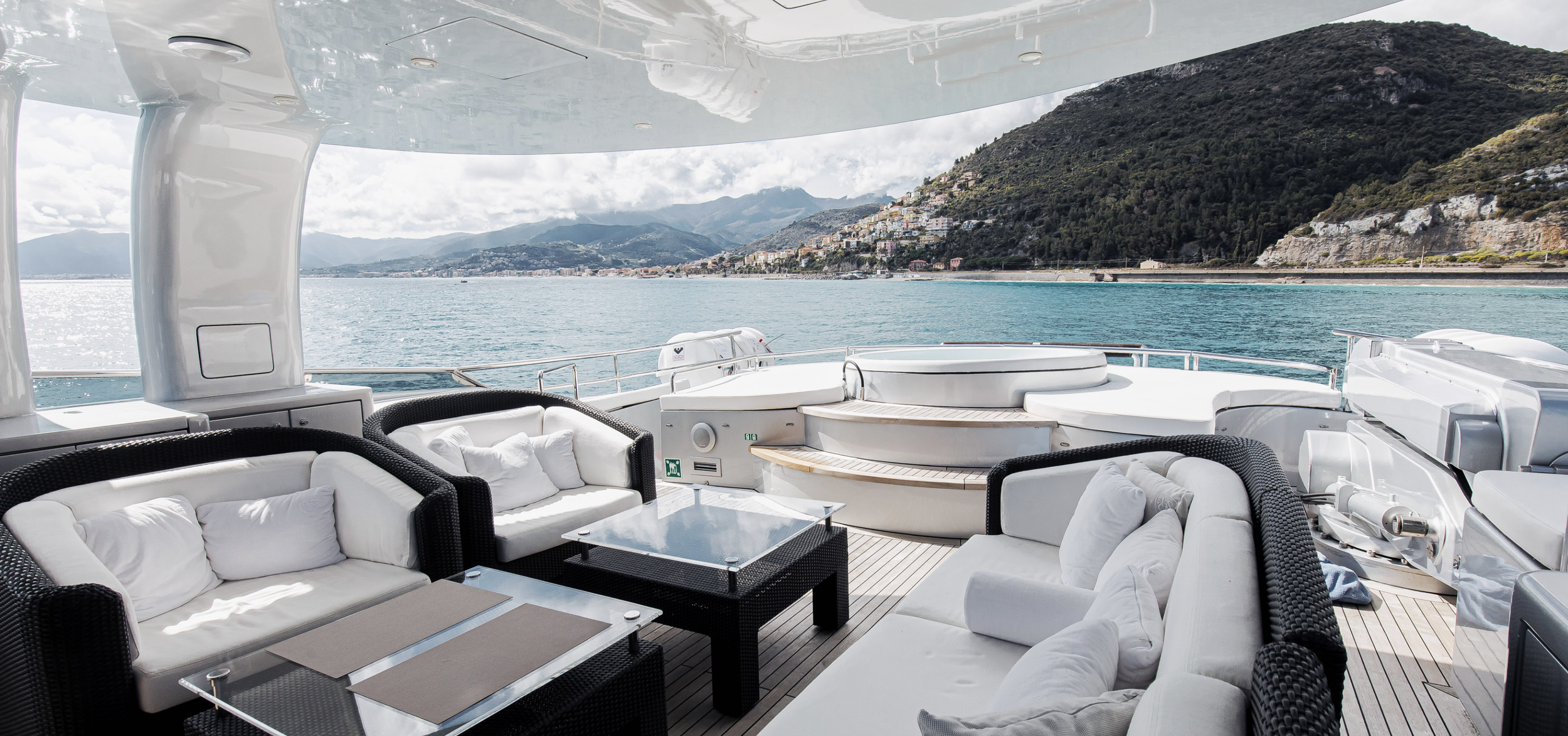 Luxury yacht deck with white seating, black wicker furniture, glass tables, and a panoramic view of the water, mountains, and town in the background.