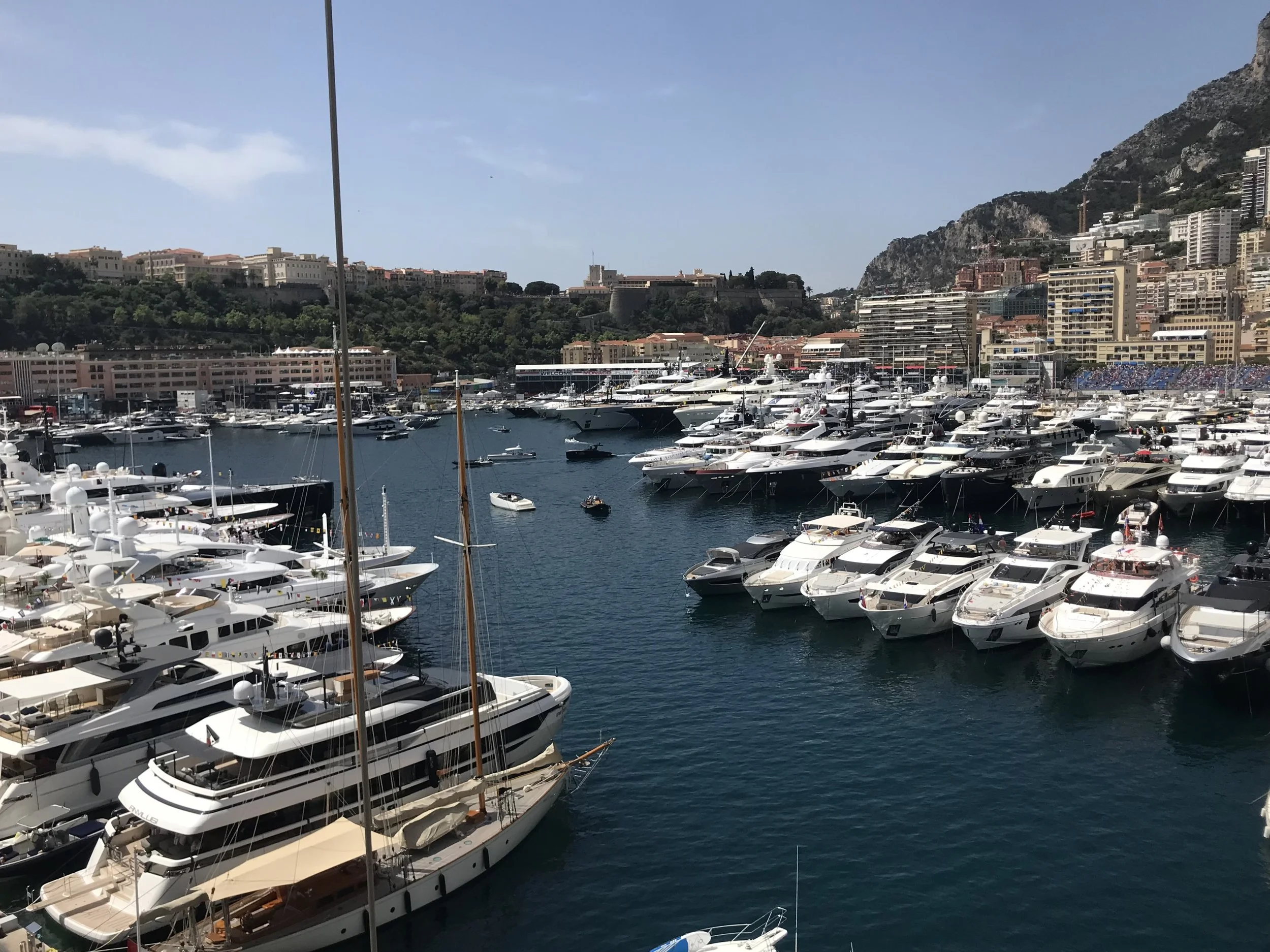 A marina filled with numerous luxury yachts and boats docked, with city buildings and green hills in the background under a partly cloudy sky.