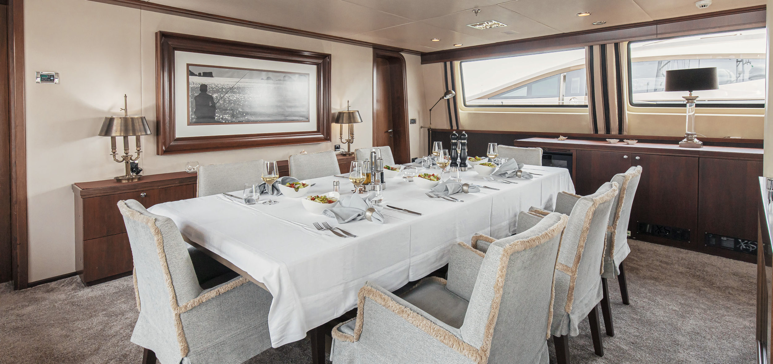 A formal dining room with a large white tablecloth-covered table set with wine glasses, plates, napkins, and salads, surrounded by upholstered chairs, and decorated with picture frames and lamps, on a yacht or boat.